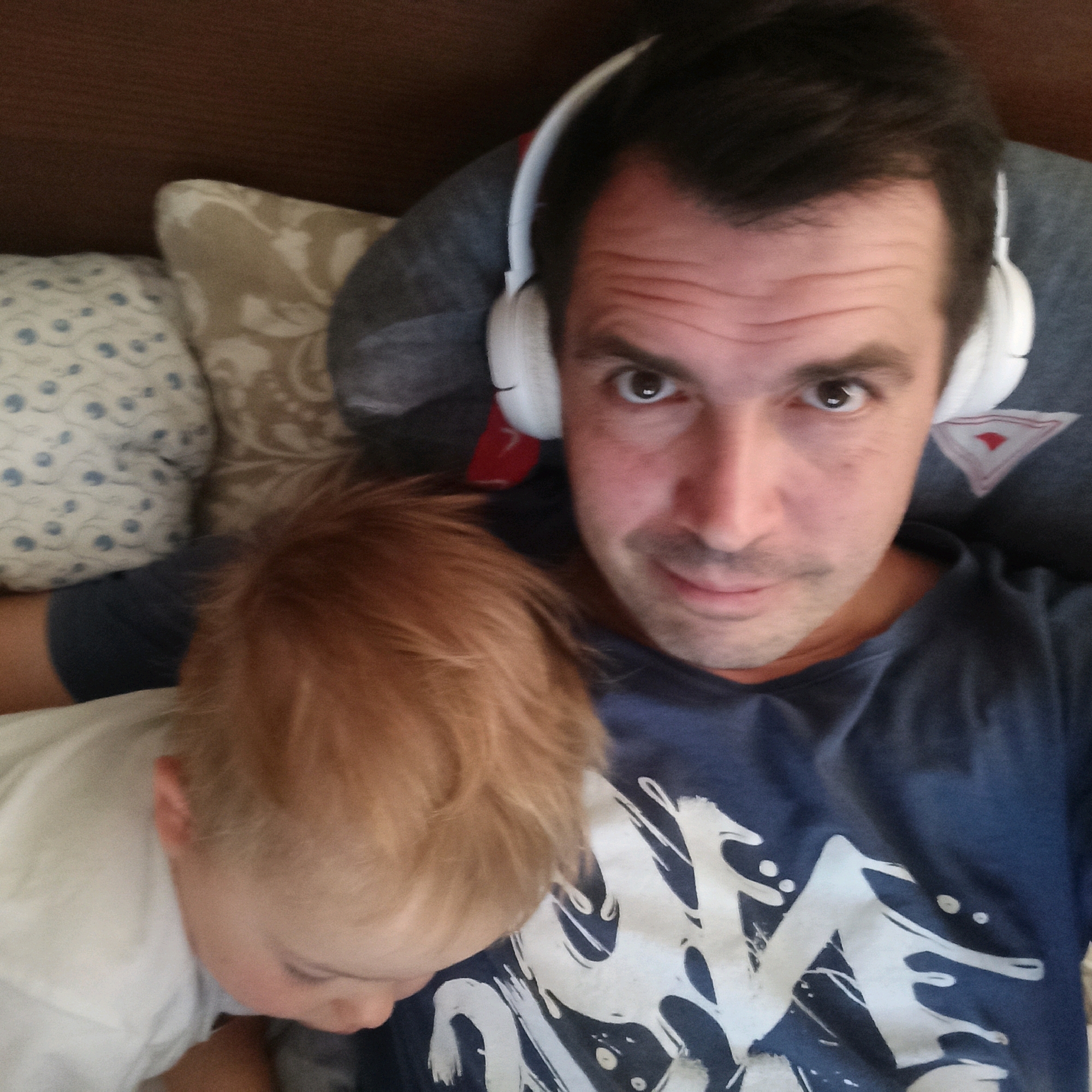 A close-up, over-the-shoulder selfie shows a man wearing white headphones. He is looking into the camera with his eyes open and a slight smile. To his left, a child with blonde hair is partially visible, looking down. Both individuals are positioned on what appears to be a bed with pillows. The man is wearing a dark blue t-shirt with a white graphic design. The environment suggests a domestic indoor setting. There is no indication of the city of Nizhny Novgorod, Russia.
