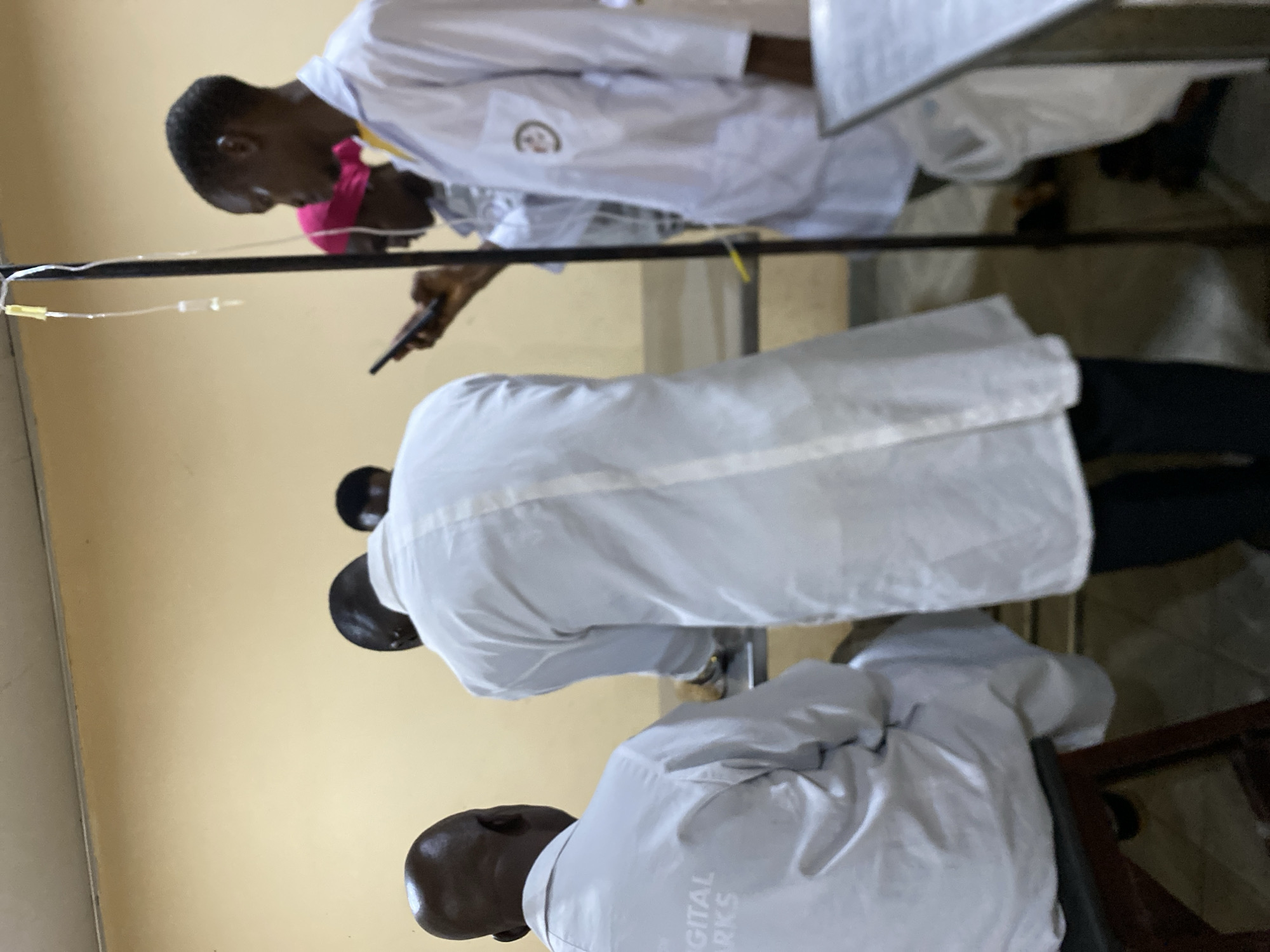 The image depicts a group of individuals, likely healthcare professionals, indoors. They appear to be examining or working on patients. All wear white coats, while one person is wearing a pink head covering. In the top left corner is an IV drip. One of the individuals in the background is holding a phone. "DIGITAL WORKS" is printed on the back of a coat. The location is in Bukuru, Nigeria.