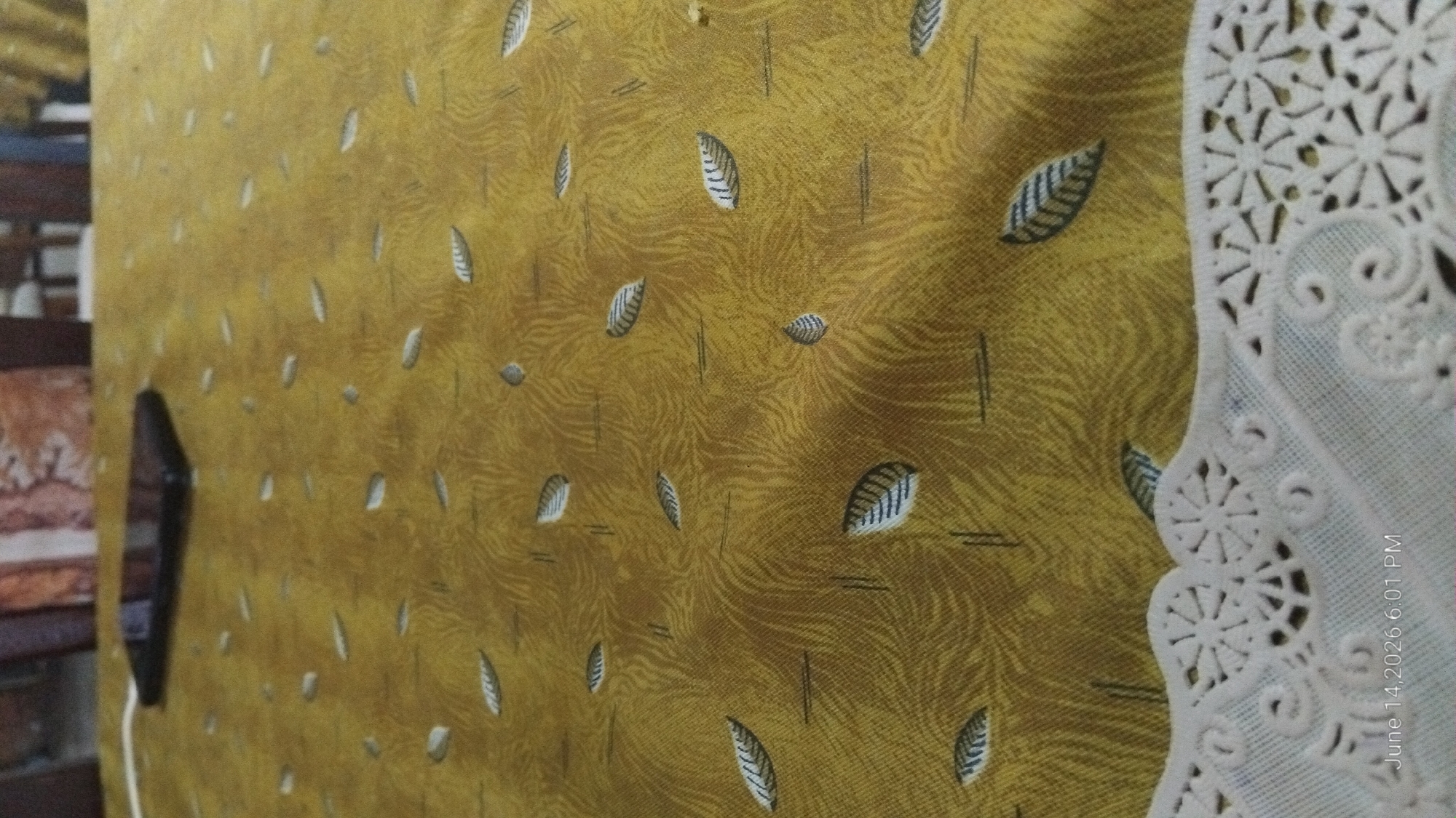 A smartphone rests on a gold-colored tablecloth with a repeating leaf pattern. The tablecloth has a textured, somewhat abstract design of flowing lines in a lighter shade of gold, interspersed with dark lines and leaf motifs. In the foreground, a white lace trim with intricate floral designs borders the tablecloth. The background is blurred but suggests the presence of chairs and furniture, implying an indoor setting. A timestamp in the bottom left corner reads "June 14, 2026 6:01 PM". This scene is likely situated within a private residence in Howayi, Nigeria, given the specific location requested. There are no people or any discernible actions or interactions occurring in the image.
