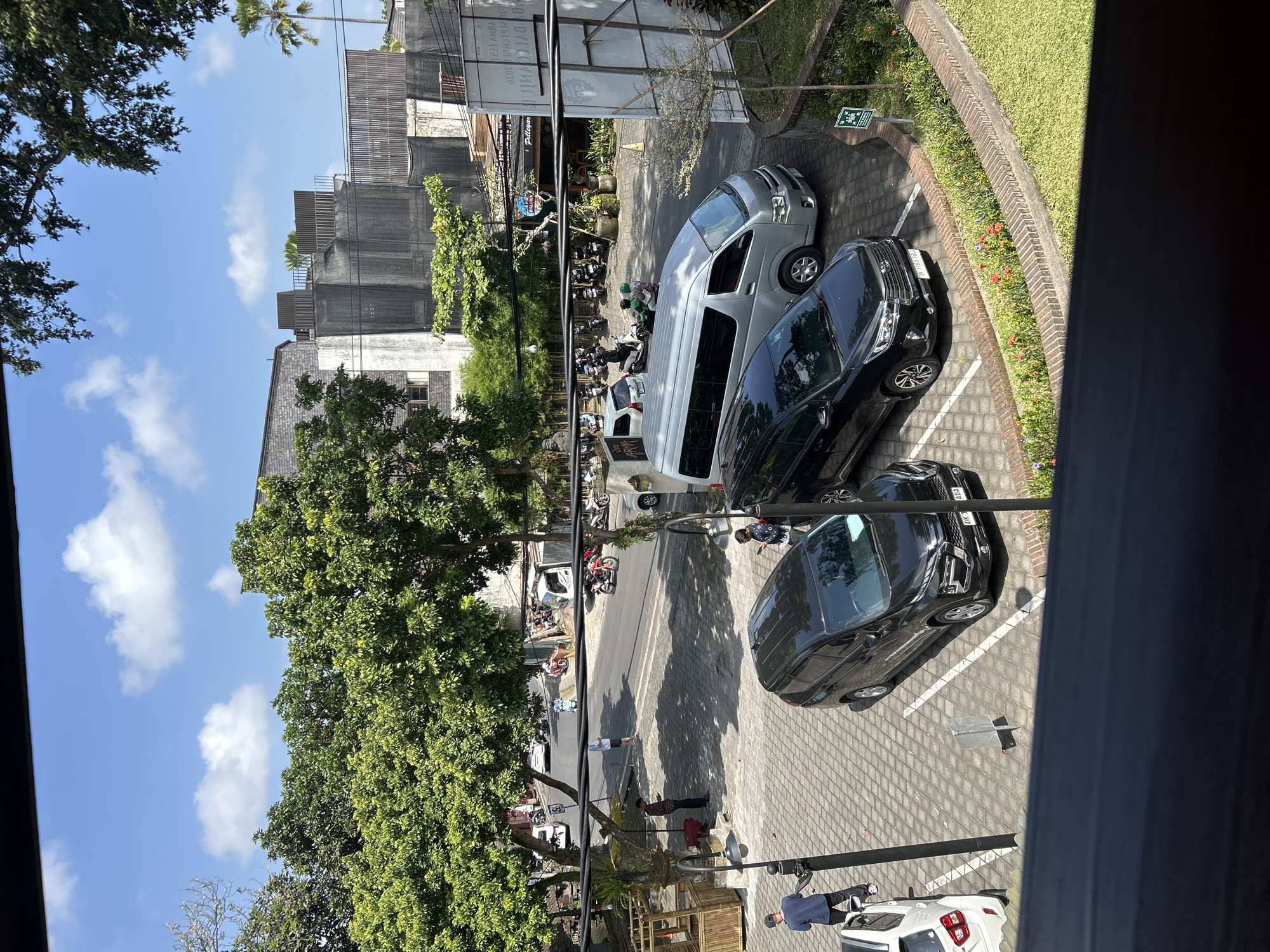 An overhead view captures a paved parking area in Ubud, Indonesia, under a bright, sunny sky with scattered white clouds. Several vehicles are parked, including two black SUVs, a silver minivan, and a white compact car. In the background, a street bustles with activity, featuring numerous motorcycles, a few cars, and pedestrians. Trees with dense green foliage line the street and surround the parking area, casting dappled shadows on the pavement. Buildings, some with modern architectural elements and others with traditional Balinese design influences, are visible behind the trees. The foreground is partially obscured by a dark, blurred railing or ledge, suggesting the image was taken from an elevated vantage point.