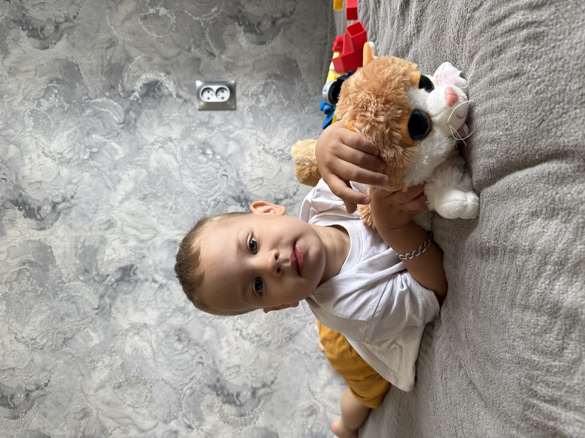 A toddler is holding a plush cat toy while lying on a gray textured surface, likely a bed or couch. The toddler wears a white shirt and yellow shorts, and a silver bracelet is visible on their wrist. The background includes a gray and white patterned wall with a double electrical outlet. The presence of the outlet and the wall suggests this scene is indoors, possibly a residence. Rusanovo, Russia is not visually confirmed by image features.