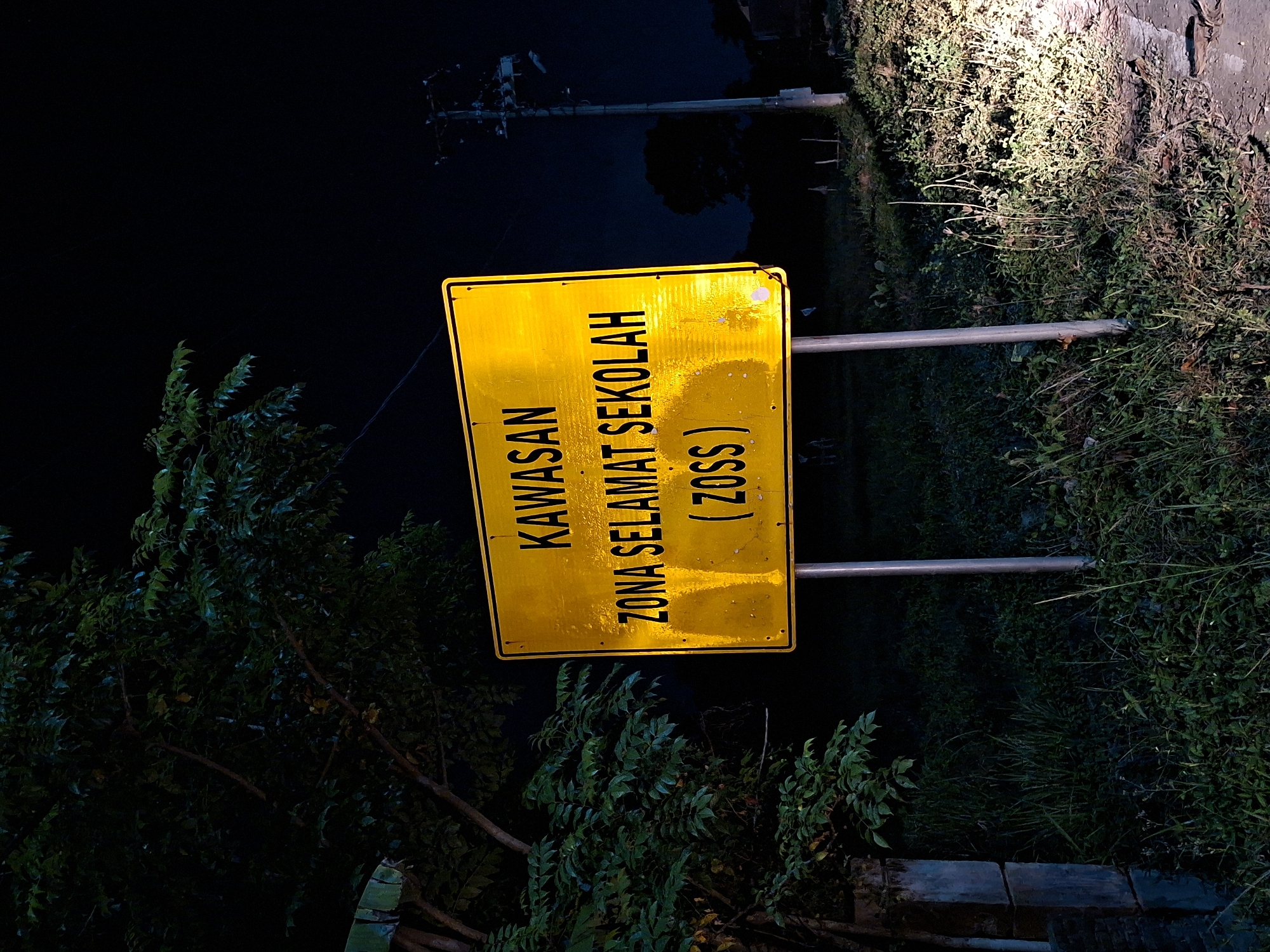 A photograph taken at night in Kutaglumpang, Indonesia, shows a yellow road sign that reads "KAWASAN ZONA SELAMAT SEKOLAH (ZOSS)". The sign is lit by an unseen light source, possibly headlights, and is positioned near vegetation. A utility pole is visible in the background. The setting suggests the sign is located near a road or path leading to or from a school.