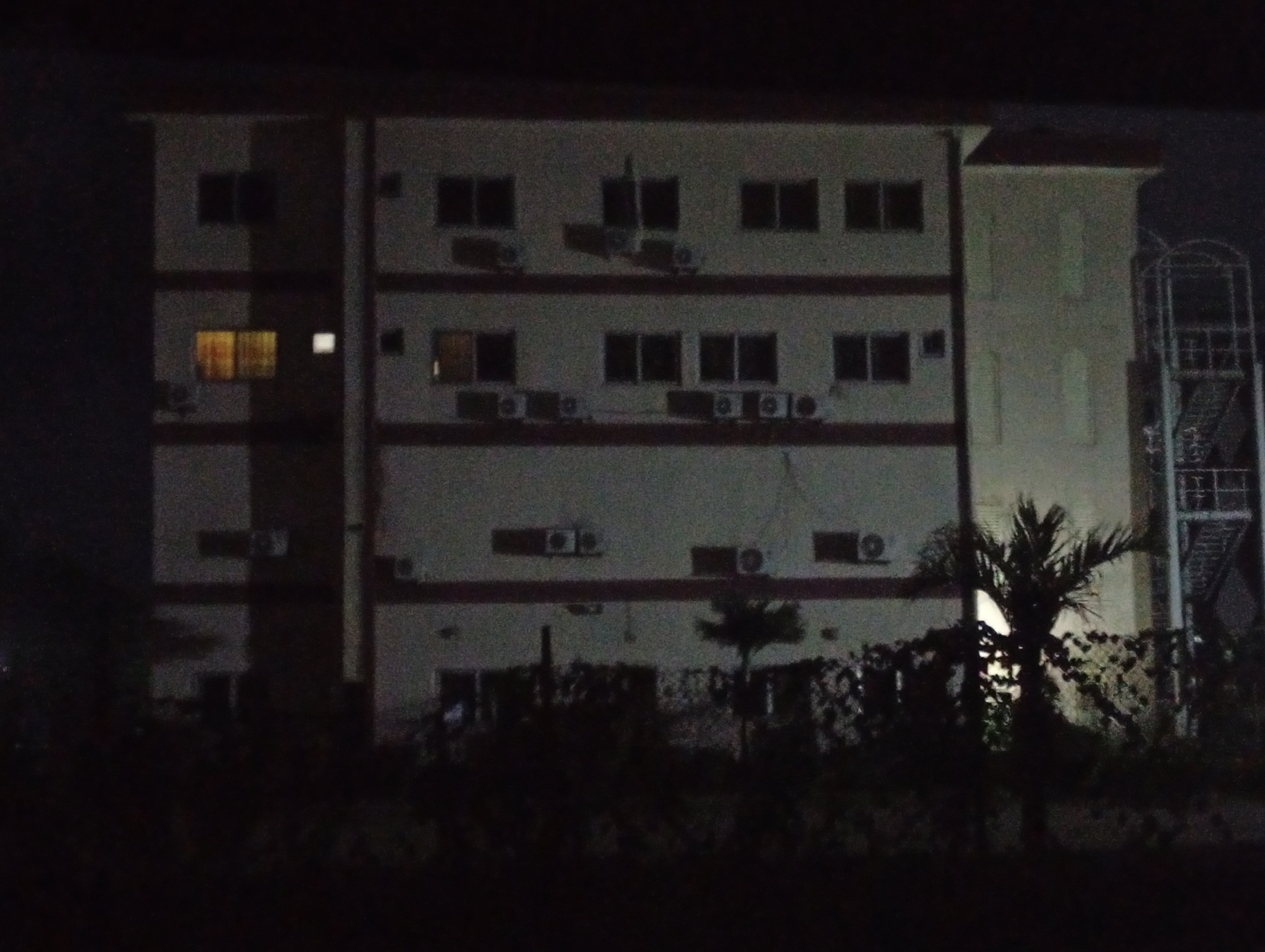 A nighttime photograph shows a building in Ijanikin, Nigeria. The building appears to be a multi-story residential or commercial structure, with several windows and air conditioning units visible on the exterior walls. A fire escape is present on the right side of the building. In the foreground, there are trees and bushes obscuring the lower portion of the building. The lighting is dim, making it difficult to discern specific details of the building's facade or the surrounding environment.
