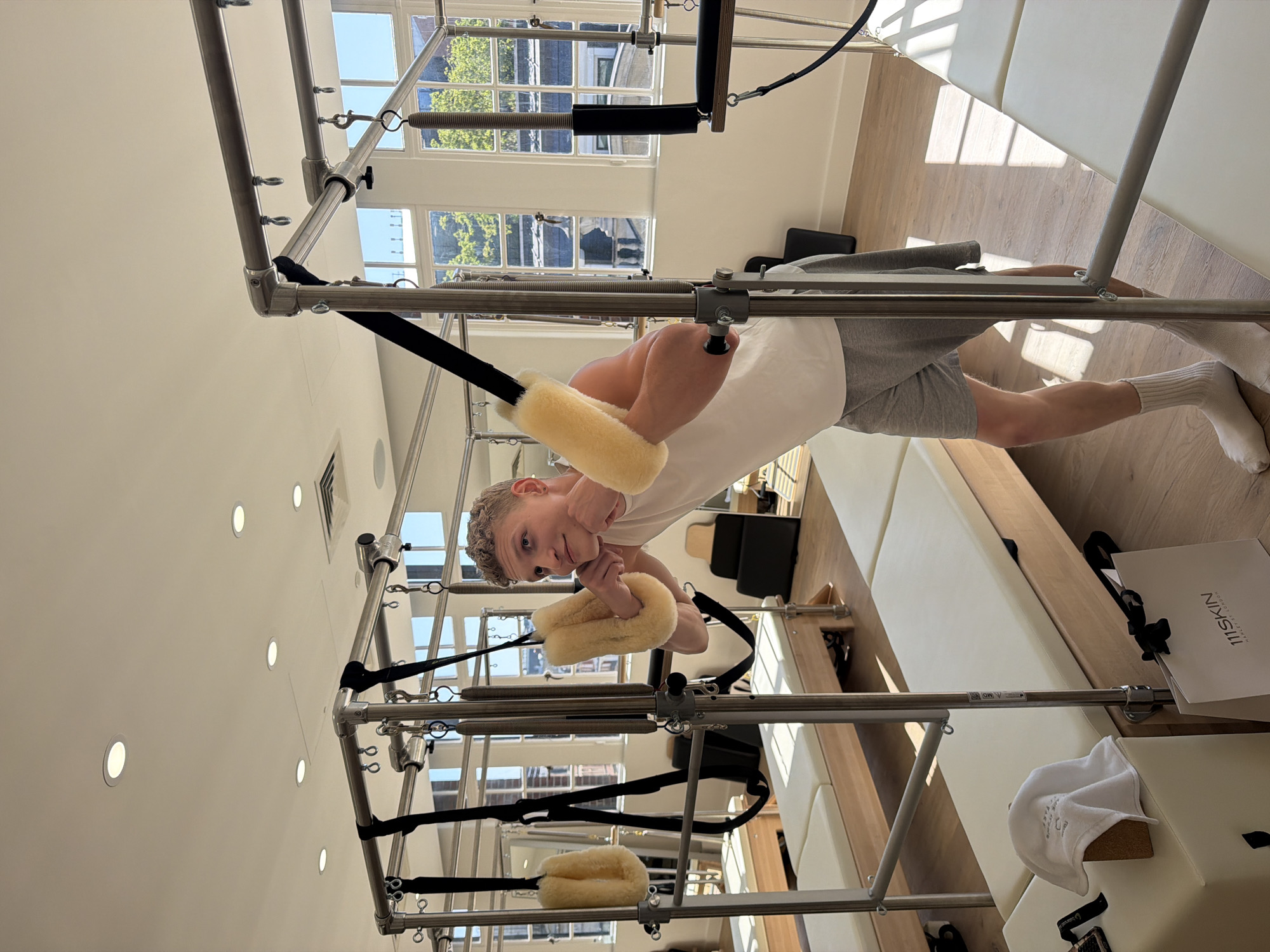 A young, light-skinned male is standing on a reformer pilates machine in what appears to be a pilates studio. The studio is well lit with natural light entering through multi-paned windows. The man is wearing a white t-shirt, grey shorts, and white socks. He is holding the reformer's padded straps with both hands, his body positioned as if he is in the middle of a pilates exercise. The machine has a chrome frame, black straps and springs. The room has light wood floors and white walls. A shopping bag with "111SKIN" is visible in the background. The location is suggested to be London, United Kingdom.