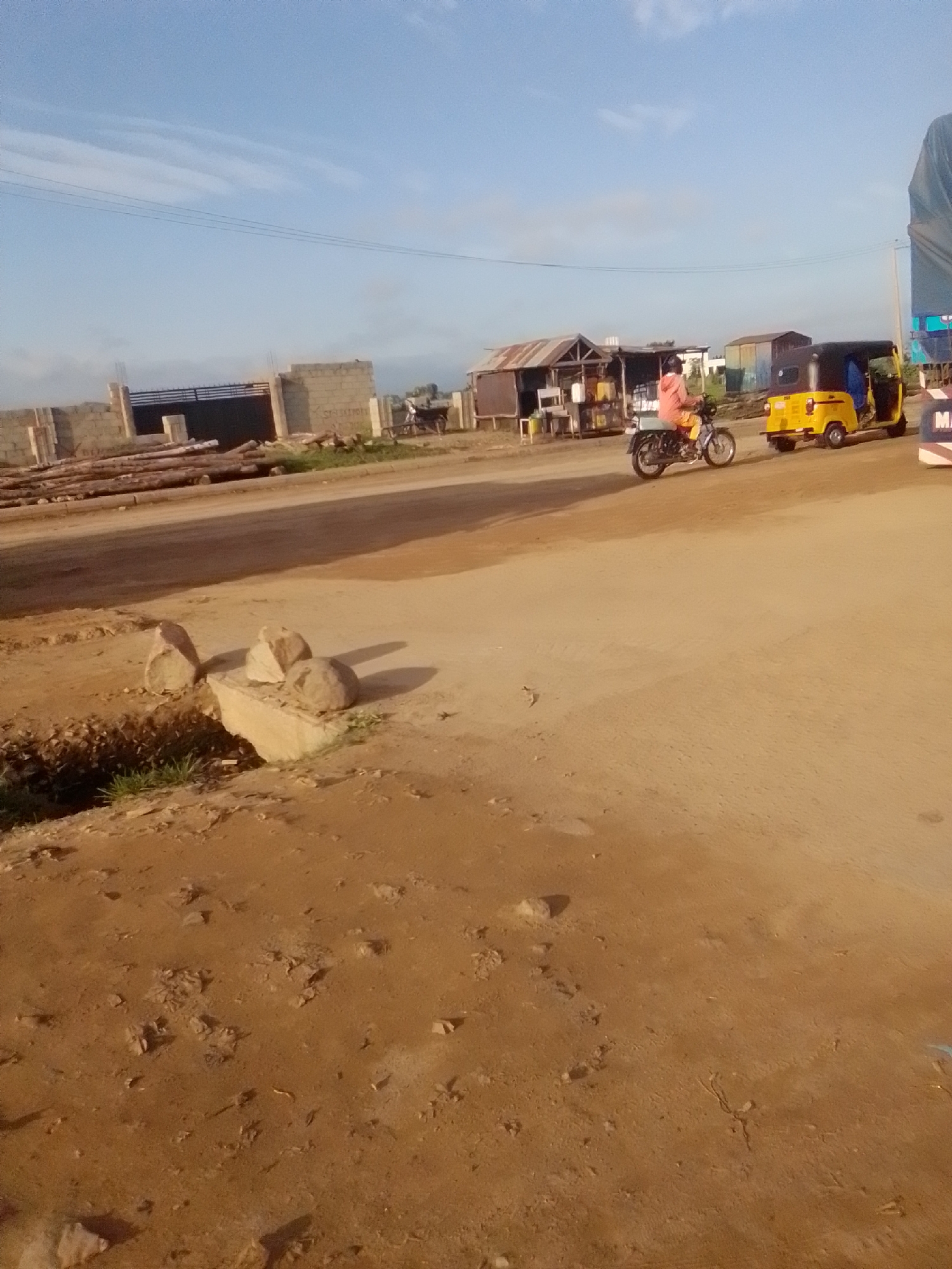 In Kwadon, Nigeria, a scene unfolds on a dusty road under a bright, partly cloudy sky. A person wearing a pink hoodie rides a motorcycle towards the right side of the frame. Beside the motorcycle, a yellow three-wheeled auto-rickshaw is parked, also facing right, with its door open. In the background, a small market stall with a corrugated iron roof displays various items, and a construction site with cinder block walls and stacked lumber is visible. The foreground of the image is dominated by the dirt road and a small drainage area with grass and several large stones resting on concrete blocks.