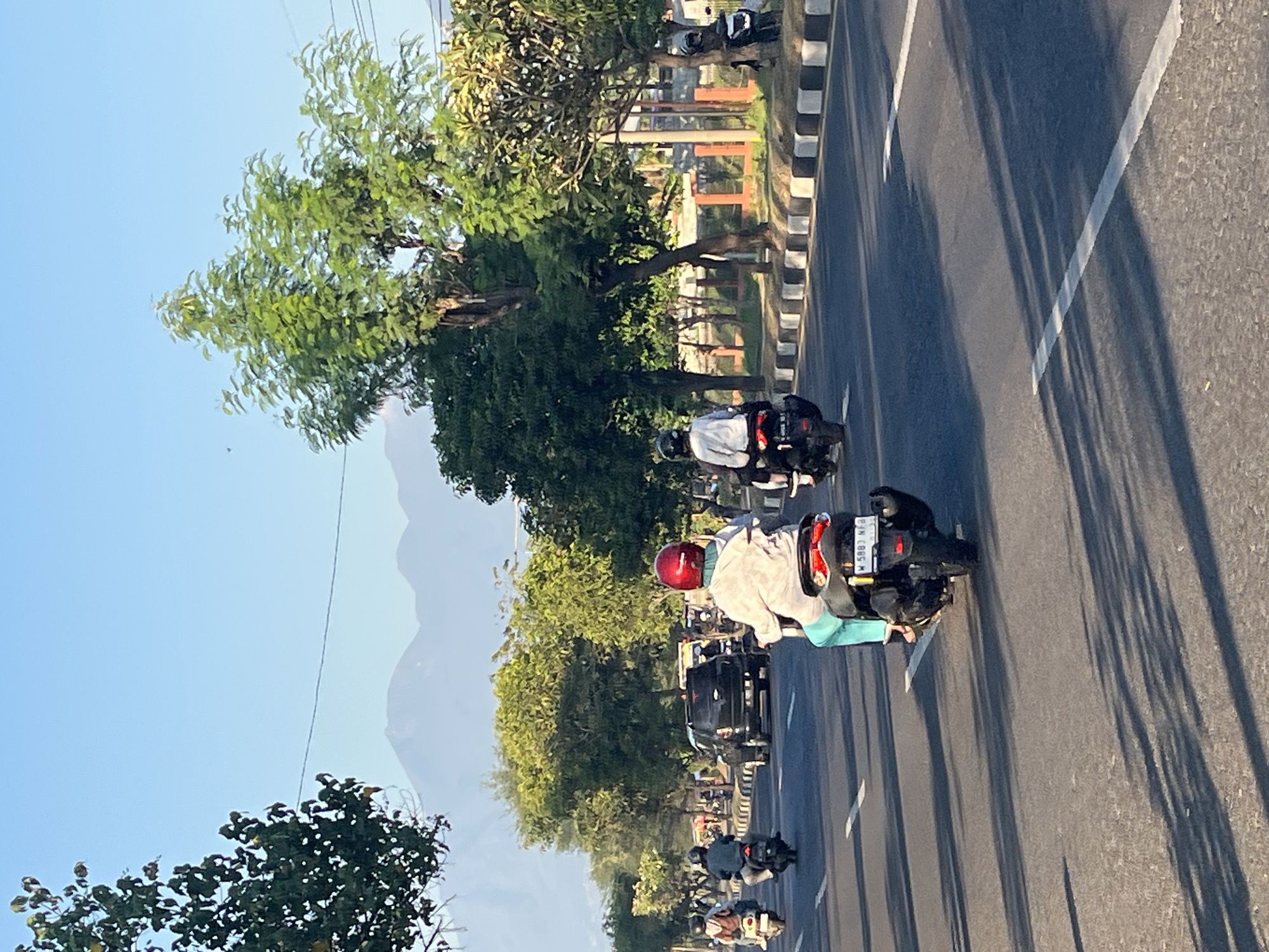 The image depicts a street scene in Siring, Indonesia, under a clear blue sky. In the foreground, several people are riding motorcycles along an asphalt road. One individual in a red helmet and a light-colored shirt with turquoise pants is prominent, riding a black motorcycle with a license plate reading "W 5883 NFB". Other motorcyclists are visible ahead, moving in the same direction. A black SUV is also on the road, with other vehicles further in the distance. Trees line the side of the road, casting long shadows across the pavement. In the background, mountains are faintly visible through a haze. The scene suggests a typical day of traffic in a location with natural landscapes.