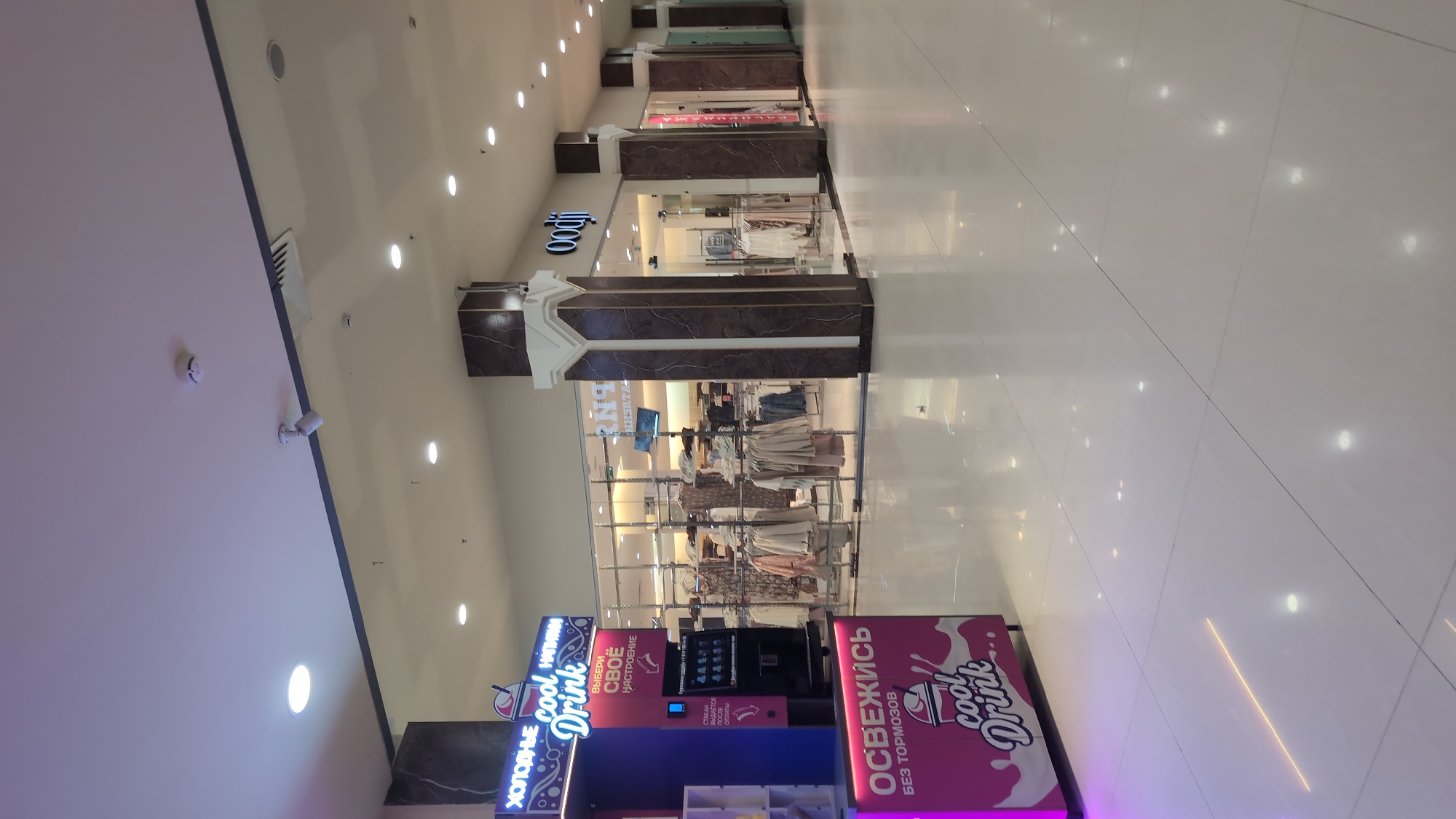 The image captures a scene within a modern shopping mall, likely in Nizhny Novgorod, Russia, given the presence of Russian text. The overall lighting is bright and artificial, typical of an indoor retail environment, with numerous ceiling lights reflected on the polished floor.
In the foreground, to the left, is a vibrant pink and purple kiosk advertising "cool Drink" with Russian text including "ОСВЕЖИСЬ" (refresh yourself) and "БЕЗ ТОРМОЗОВ" (without brakes). Next to it, on the left side of the frame, is a larger display with neon blue lettering that reads "ХОЛОДНЫЕ COOL Drink НАПИТКИ" (cold cool drink beverages). This display features a stylized graphic of a milkshake with a straw. Below the "cool Drink" text, there's a section with Russian text that translates to "CHOOSE YOUR MOOD." A black vending machine with a touch screen is integrated into this kiosk, with instructions in Russian below it indicating to select and then pay.
Further into the mall, there are several storefronts. The most prominent one in the mid-ground is branded with "oodji" in white lettering against a dark background. This store has large glass windows displaying clothing on racks, suggesting it is a fashion retailer. The store's interior is well-lit, and mannequins are visible inside. The storefronts are separated by large, decorative pillars with a marble-like pattern in shades of brown and beige. The floor is a continuous expanse of light-colored tiles with a high-gloss finish, reflecting the lights and creating a sense of spaciousness. The ceiling is high and features a mix of recessed lighting and ventilation grilles. No people are visible in the image.