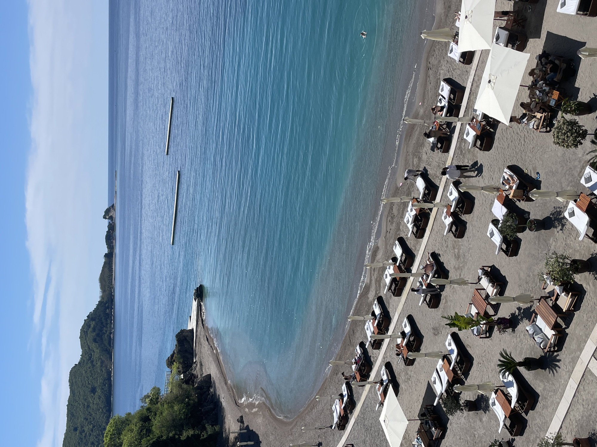 The image shows a beach on a sunny day in Budva, Montenegro. The beach is lined with lounge chairs and umbrellas, some of which are occupied by people. The water is calm and clear, with a few floating safety barriers. On the left of the image, the mountainous coastline curves to form the edge of the bay. The image is taken from a high angle, likely from a building or elevated position.