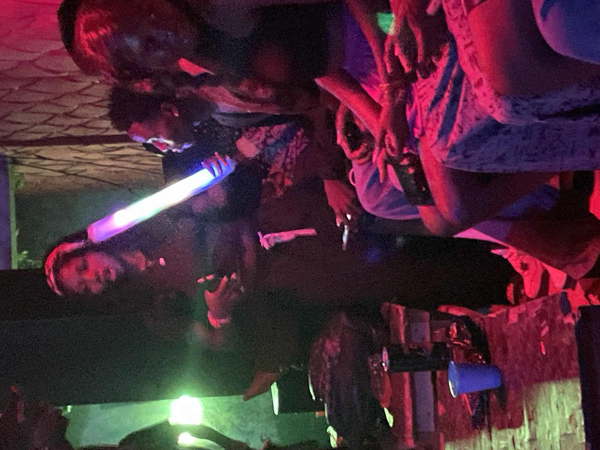 The image depicts an indoor scene, likely a club or party, in Durumi, Nigeria. Several individuals, appearing to be of African descent, are gathered. In the foreground, a woman wearing a headband is holding a glowing, multi-colored tube. To her right, another individual is visible, and further to the right, a group of people are seated, some wearing purple clothing. The environment is dimly lit and illuminated by colored lights, primarily red and green. Various objects, including a blue cup and what appear to be speakers, are present in the scene. The overall atmosphere suggests a social gathering or event.