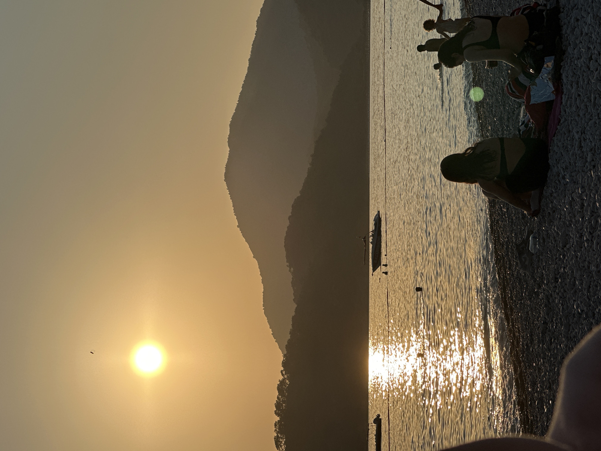 This image depicts a sunset over a bay in Fethiye, Turkey. The sun, a bright white orb, is positioned in the upper-left sky, casting a strong golden-orange glow across the hazy sky. A small, dark, elongated object is visible in the sky above the sun. The background features multiple layers of mountains, appearing as dark, hazy silhouettes, with the closest slopes covered in dense vegetation. The body of water, likely the sea, reflects the sun's light in a prominent, shimmering golden path extending towards the viewer. A red buoy line stretches horizontally across the water in the mid-ground.
Two boats are present on the water: a small, dark inflatable vessel is partially visible on the far left, and a larger white motorboat, labeled "EXCURSION" on its side, is positioned towards the right. Several individuals are visible as dark silhouettes in the water; two are near the white motorboat, and three others, including a child, are further to the right.
The immediate foreground consists of a pebble beach. Two women are seated on the pebbles with their backs to the camera. The woman on the left is wearing a dark swimsuit. The woman on the right, also in a dark top, is seated on a striped blanket, with a white item (possibly a bag) beside her. A discarded plastic bottle is visible on the pebbles to the right of the seated women. Part of a person's shoulder and arm is visible in the extreme lower-left foreground. A circular green lens flare is present over the water in the lower-right quadrant.