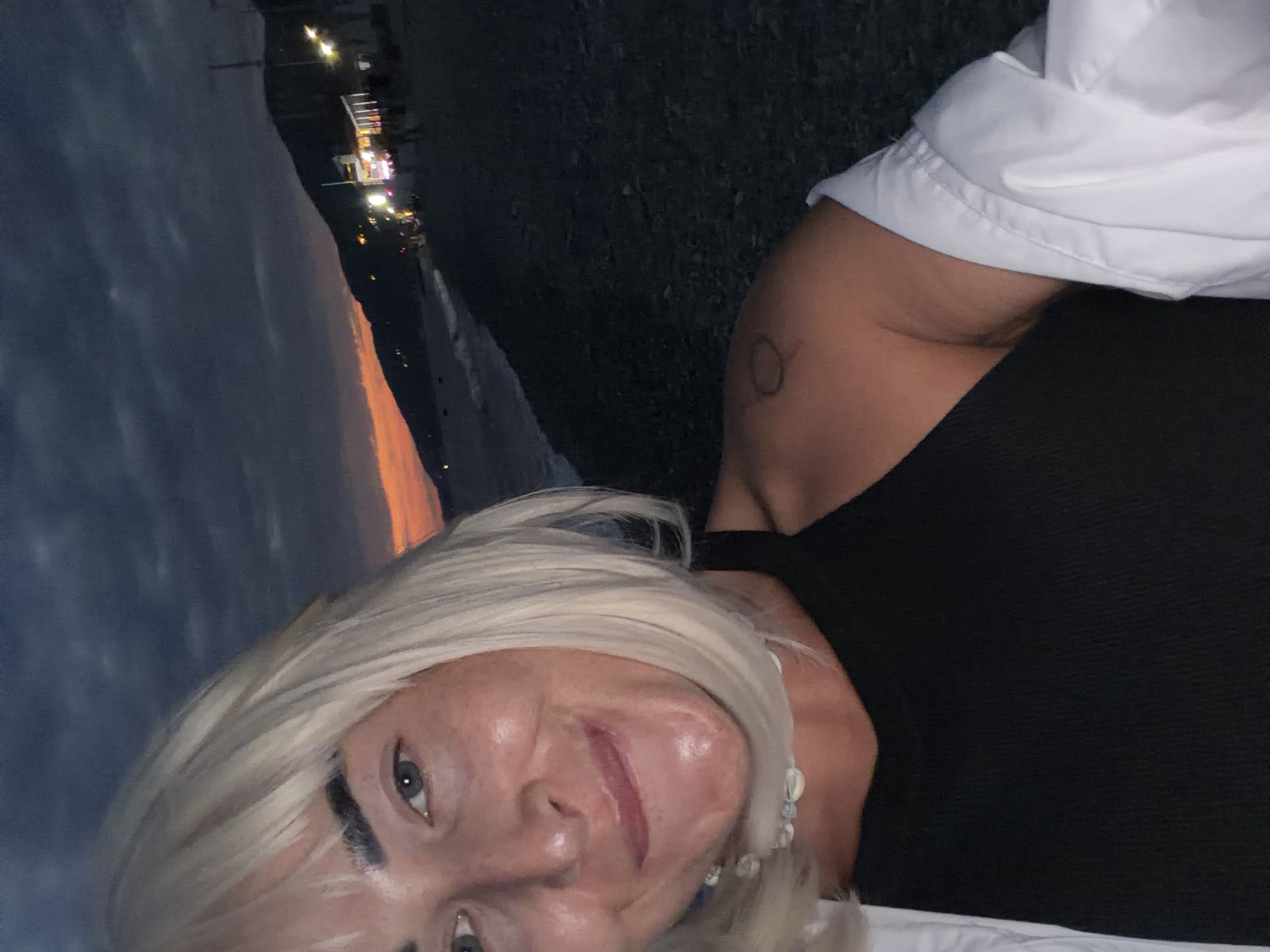 A woman with blonde hair is taking a selfie on a beach at dusk. She is wearing a black tank top and a white shirt draped over her shoulders. In the background, there are lights from buildings along the coast and the sky is a dark blue with hints of orange and pink from the sunset. The beach appears to be made of pebbles. The location is likely Sochi, Russia, as indicated by the context.