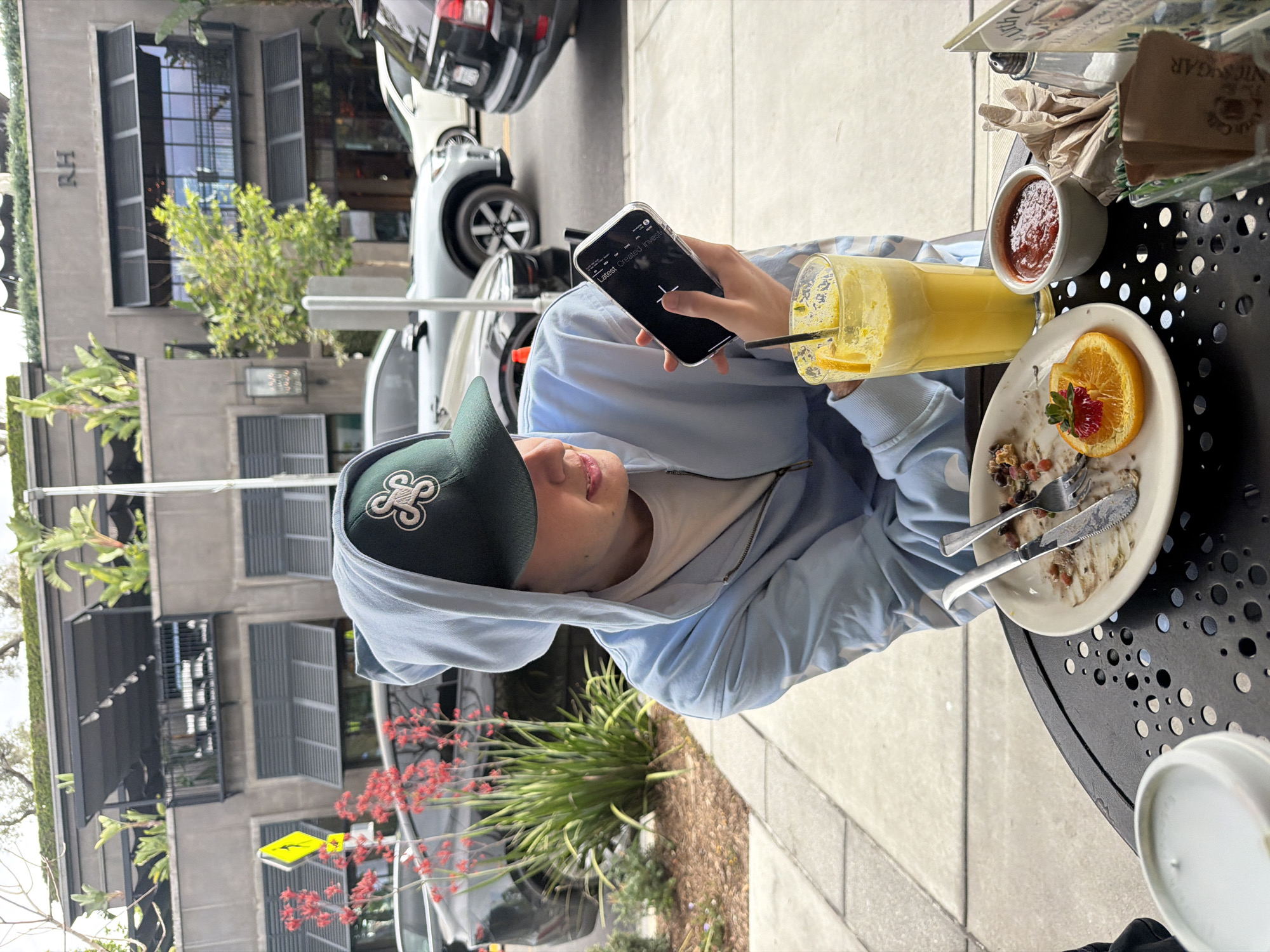 Here is a description of the image:
An outdoor shot of a person seated at a round, black metal table. The person, wearing a light blue hooded sweatshirt and a dark green baseball cap with a white emblem, is holding a smartphone with a black screen in their left hand. On the table, there's a tall glass of orange juice with a straw, a small bowl containing a reddish sauce, and a partially eaten dish garnished with an orange slice and a strawberry. A knife and fork rest on the dish. Condiments and other items are also visible on the table. In the background, there is a commercial building with windows and greenery. Cars are parked along the street, and the sidewalk is visible next to the seating area.
This image was taken at RH Rooftop Restaurant in West Hollywood, United States of America.