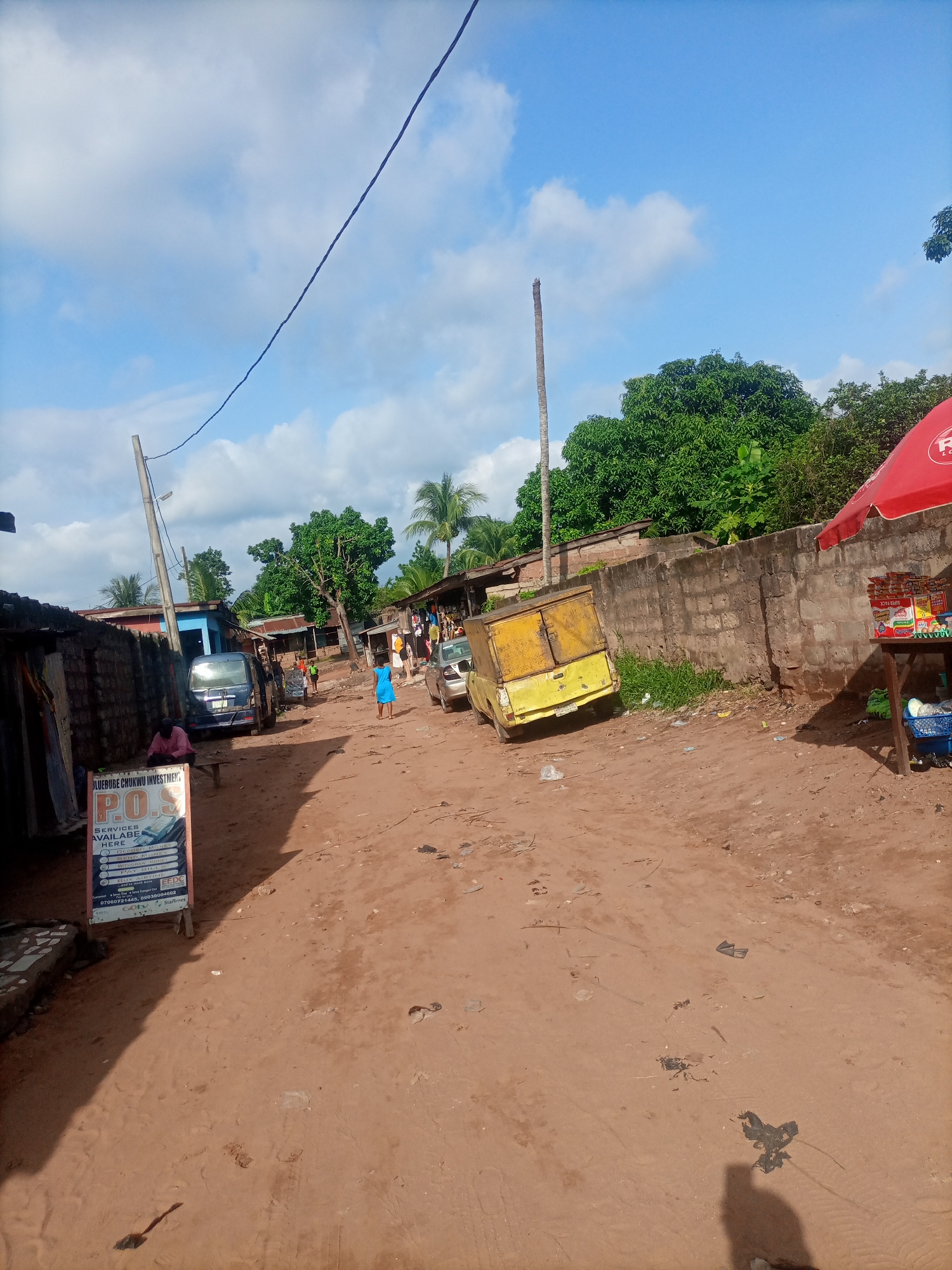 A dusty, unpaved street in Nkpor, Nigeria, is depicted under a bright, cloudy sky. The street is lined with low-rise buildings and a thick, red brick wall. Several vehicles are parked along the side of the road, including a blue van and a yellow pickup truck with a cargo box. A wooden table covered by a red umbrella displays various packaged goods for sale, with a blue crate of items underneath. A person in a blue dress walks down the street, and other figures are visible in the distance. A sign in the foreground advertises "P.O.S. SERVICES AVAILABLE HERE," listing services such as depositing and withdrawing money. Electrical wires crisscross the sky, supported by a wooden pole. The ground is covered in reddish-brown dirt, with scattered debris and footprints.