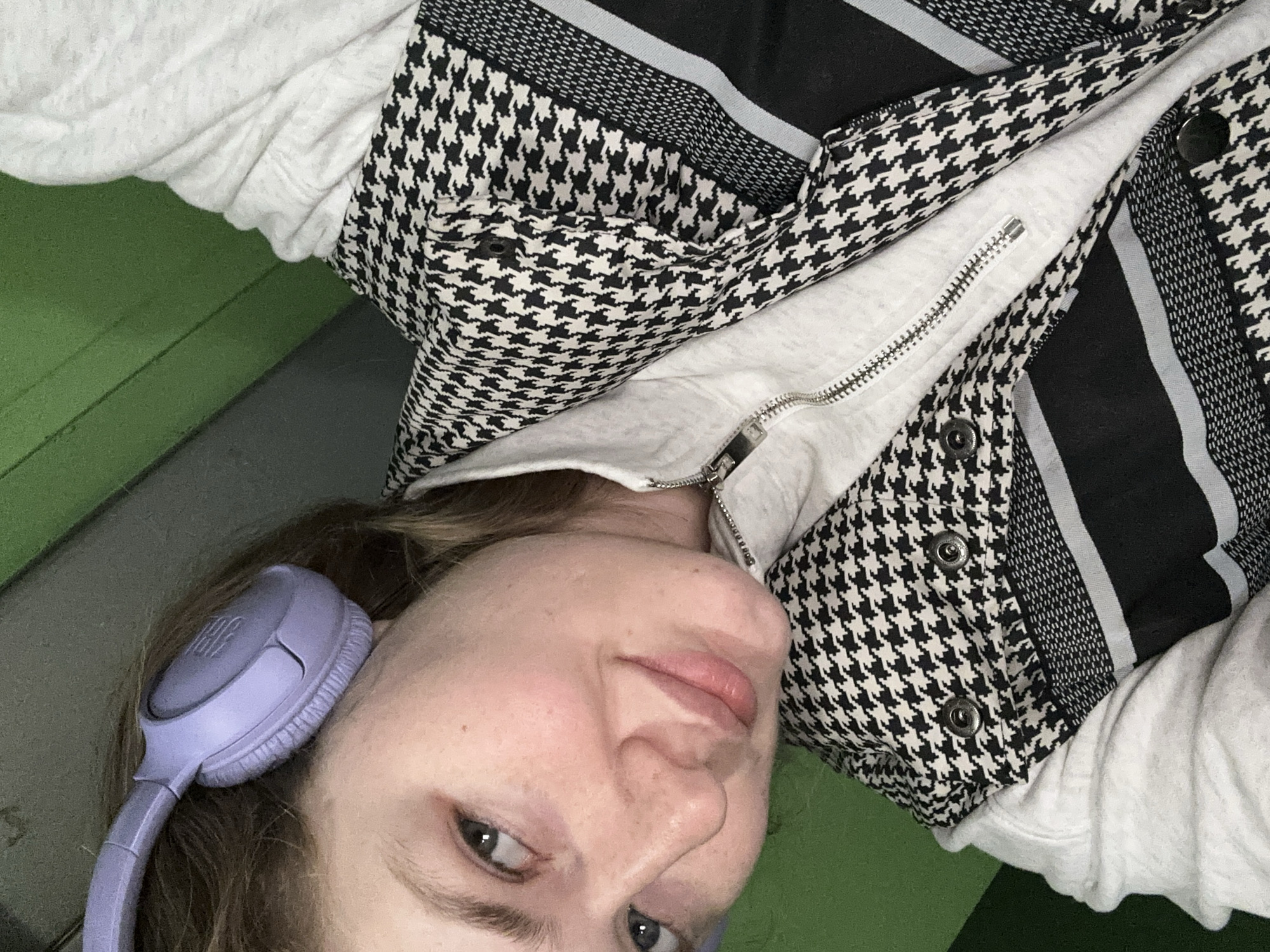 A woman with headphones is lying down in Kazan, Russia. She is wearing a white sweatshirt and a black and white houndstooth patterned vest with a zipper. The background is green and grey.