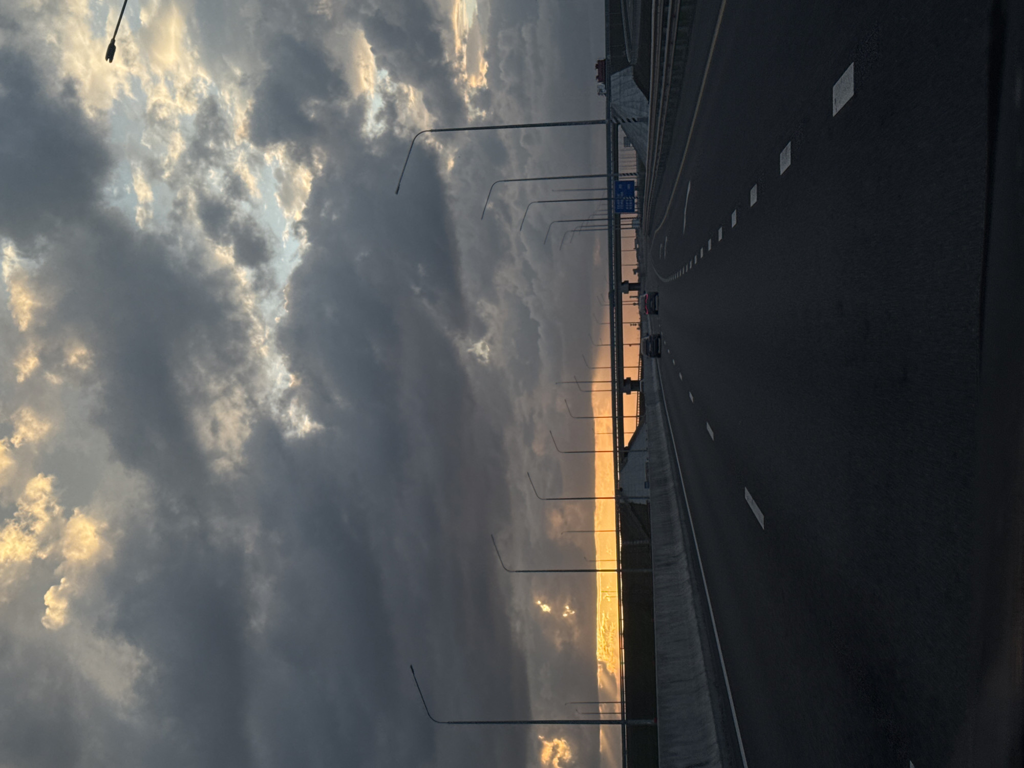 The image shows a highway in Maryanskaya, Russia, at sunset. The sky is filled with clouds, with golden light shining through. Streetlights line the side of the highway, and a few cars are visible in the distance. The road is paved and marked with white lines.
