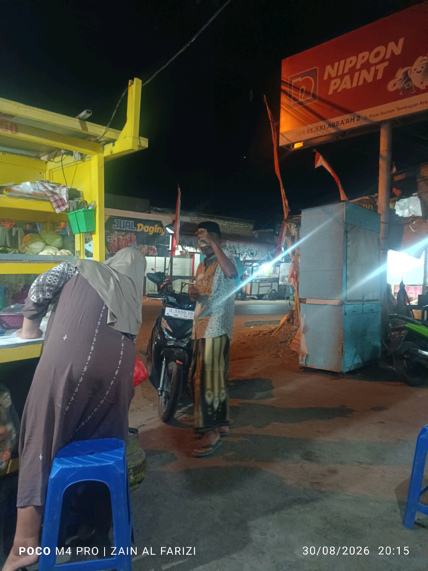 The image depicts a nighttime scene on a street in Buduran, Indonesia, likely a commercial area. A woman wearing a brown hijab and a patterned top is partially visible on the left, appearing to be attending to a food stall with a yellow awning. In the center, a man in a patterned shirt and sarong is holding a phone to his ear, possibly making or receiving a call. A motorcycle is parked behind him. To the right, there are two blue coolers or storage units. In the background, a large red billboard advertises "NIPPON PAINT" with a tagline in Indonesian, and another banner reads "JUAL DAGING" (Meat for Sale). The ground is paved, and the overall lighting suggests artificial streetlights and the illumination from the stall and billboard. The timestamp in the lower right corner indicates the photo was taken on August 30, 2026, at 20:15.