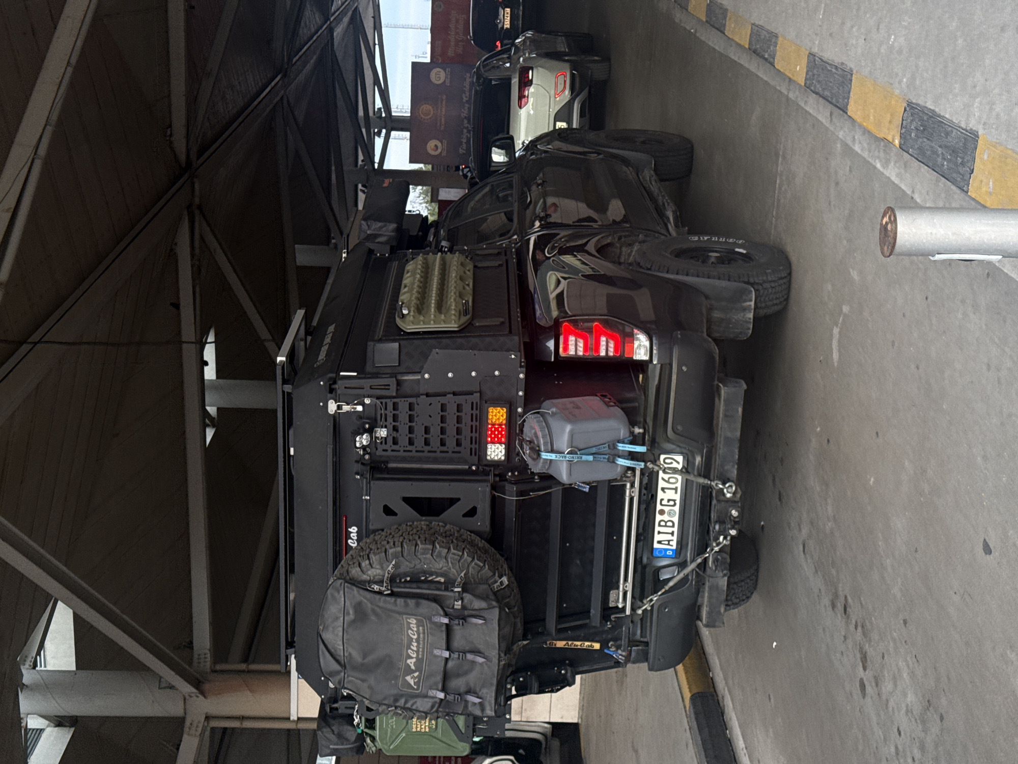 The image depicts the rear of a modified black pickup truck, possibly a Toyota Tacoma, in what appears to be a covered parking area in Edirne Merkez, Turkey. The truck has a license plate of "AIB G 1609". A spare tire is mounted on the back, along with cargo bags, water canisters, and other off-road equipment. The back of the truck is labeled "Alu-Cab". In the background, a light-colored car and a black car are visible, parked behind the truck. The area appears to be an indoor border crossing point with Turkish signage in the background: "Türkiye'ye Hoş Geldini - Hoş geldiniz".