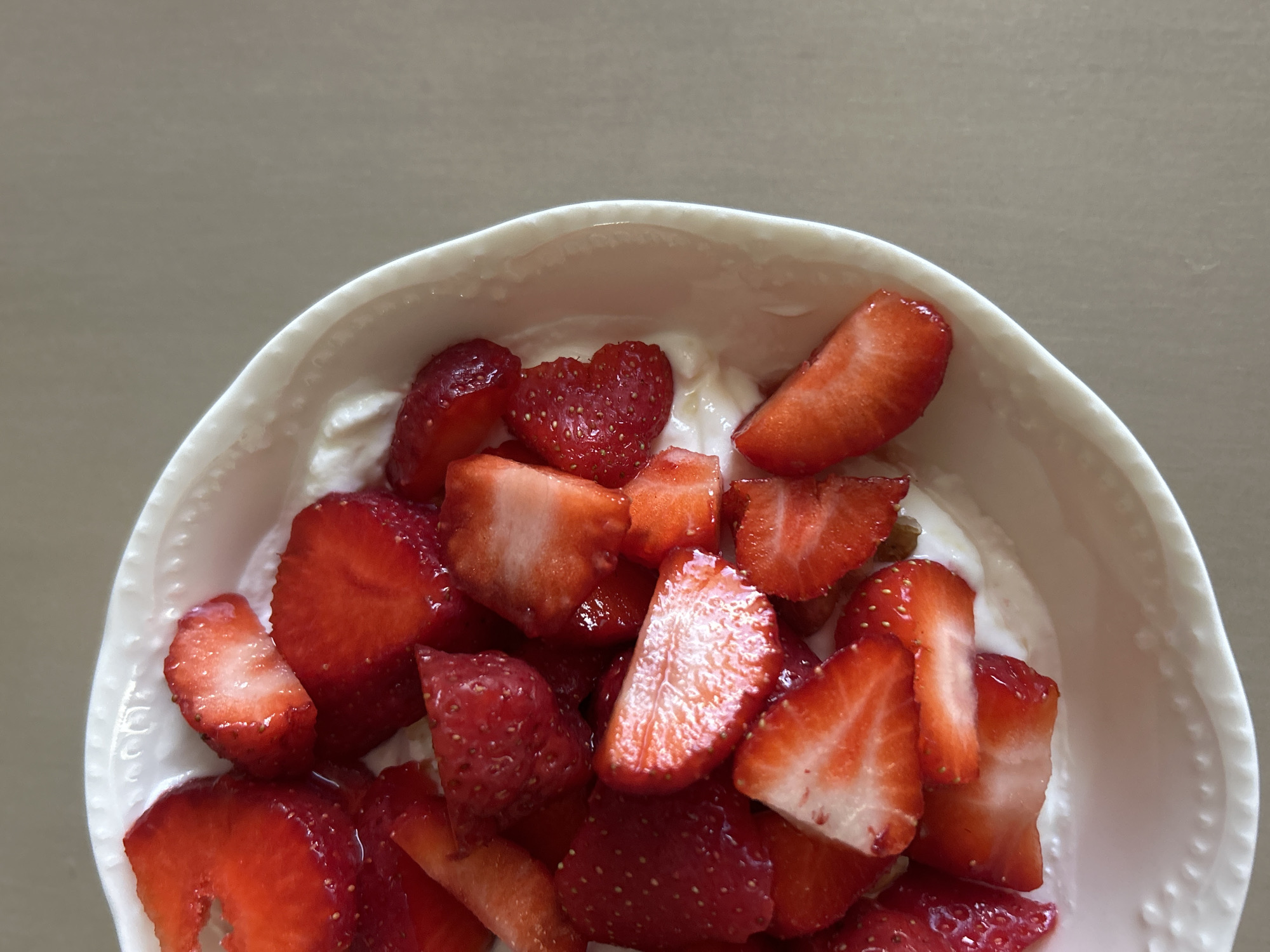 A close-up, overhead shot captures a white, textured bowl filled with yogurt and sliced strawberries. The bowl is positioned on a light brown, smooth surface, possibly a countertop or table. The strawberries, in various stages of ripeness and cut into distinct pieces, are scattered across the creamy white yogurt. Some strawberries are whole or halved, while others are cut into wedges or irregular shapes, showcasing their bright red exterior and white interior flesh. Tiny seeds are visible on the surface of the strawberry slices. The lighting is bright, creating highlights and shadows on the fruit and bowl, and the background is mostly out of focus, with the dominant color being a neutral beige. There are no people or identifiable landmarks in the image. This scene does not appear to be located in the city of Plovdiv, Bulgaria, as there are no elements that would suggest such a location.