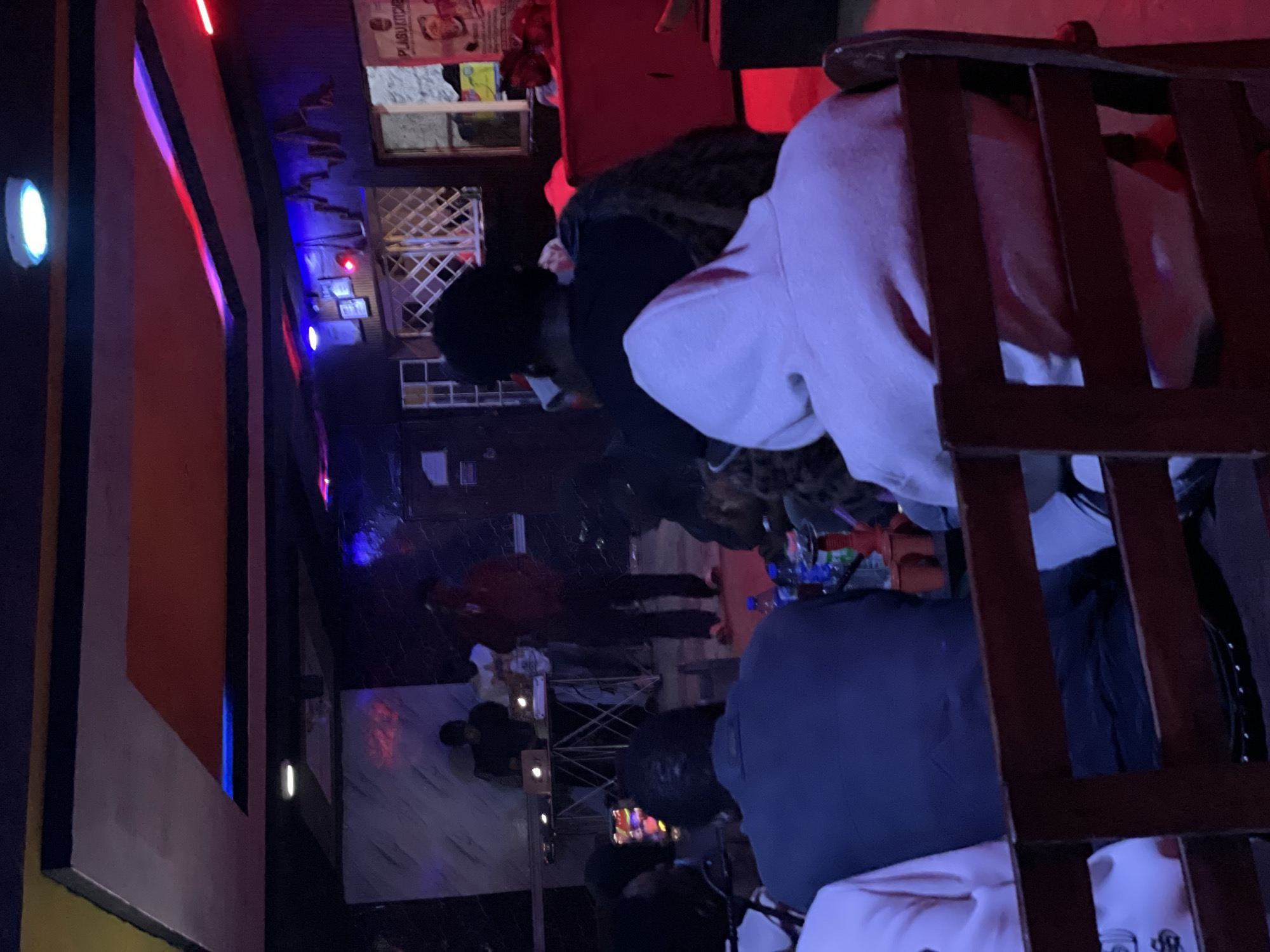 The image depicts a dimly lit indoor venue, possibly a club or lounge, in Durumi, Nigeria. The ceiling features recessed lighting with red and blue accents. In the background, three individuals are visible near a DJ booth or performance setup, with one person appearing to be speaking into a microphone. A crowd is seated in the foreground, facing away from the camera, observing the activity. The seating area is furnished with wooden chairs and benches. Red lighting illuminates parts of the scene, creating a vibrant atmosphere. Bottles of water and a hookah are present on a table in the middle ground. The overall impression is that of a social gathering or entertainment event.