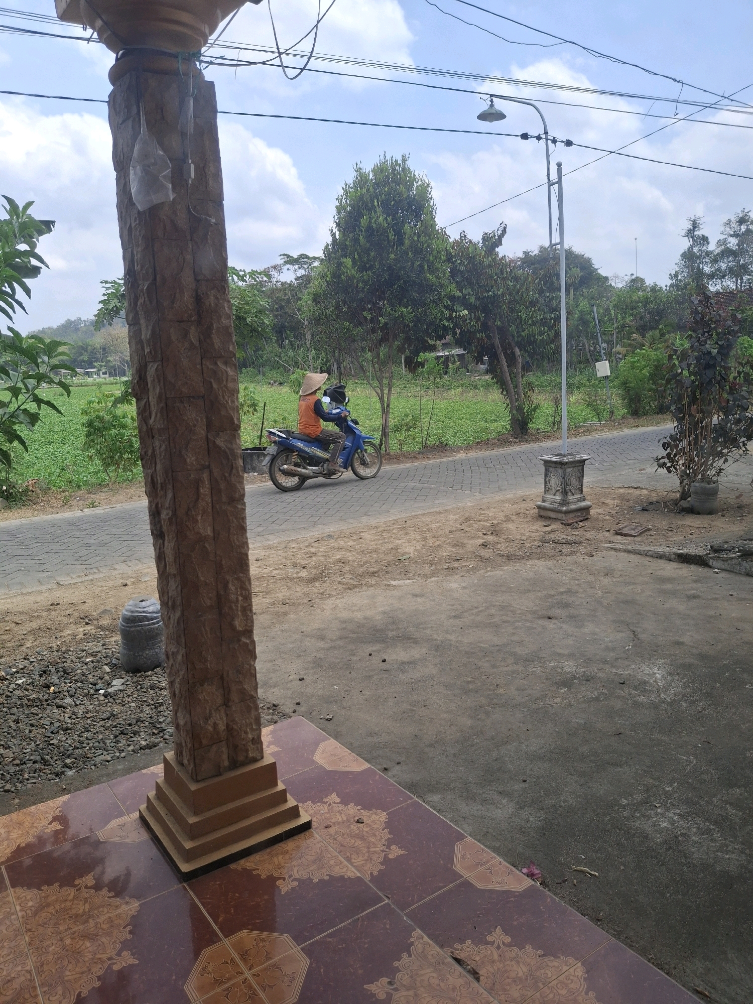 In Ngadirenggo, Indonesia, a person wearing an orange shirt and a conical hat rides a blue motorcycle down a paved road. The road is bordered by a green field on the left and a tree line on the right. A stone pillar stands in the foreground, and a street lamp is visible in the background. The sky is blue with scattered clouds.