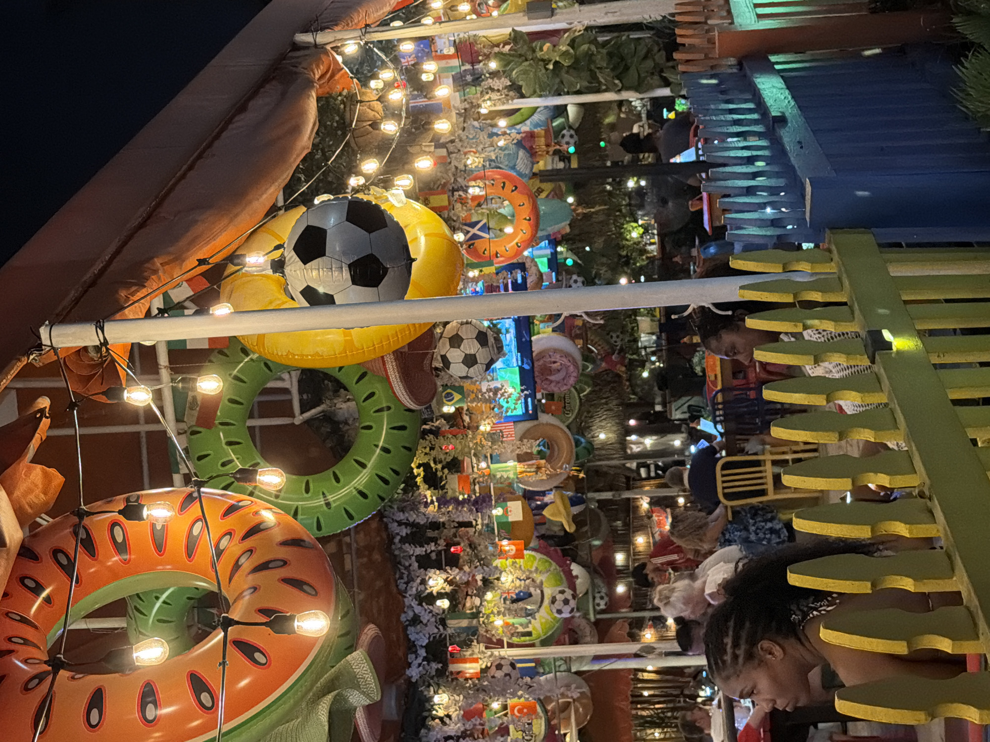This is a nighttime, eye-level, medium shot of an outdoor dining area in Hollywood, USA, decorated with inflatable pool rings, soccer balls, and string lights, suggesting a festive or sports-themed atmosphere. The foreground features a yellow and green wooden fence. Behind the fence, patrons are seated at tables, some engaged in conversation or using electronic devices. The background includes lush greenery and more decorative elements, indicating a restaurant or bar setting. The overall lighting is dim, with string lights providing warm illumination, creating a lively yet intimate ambiance.
