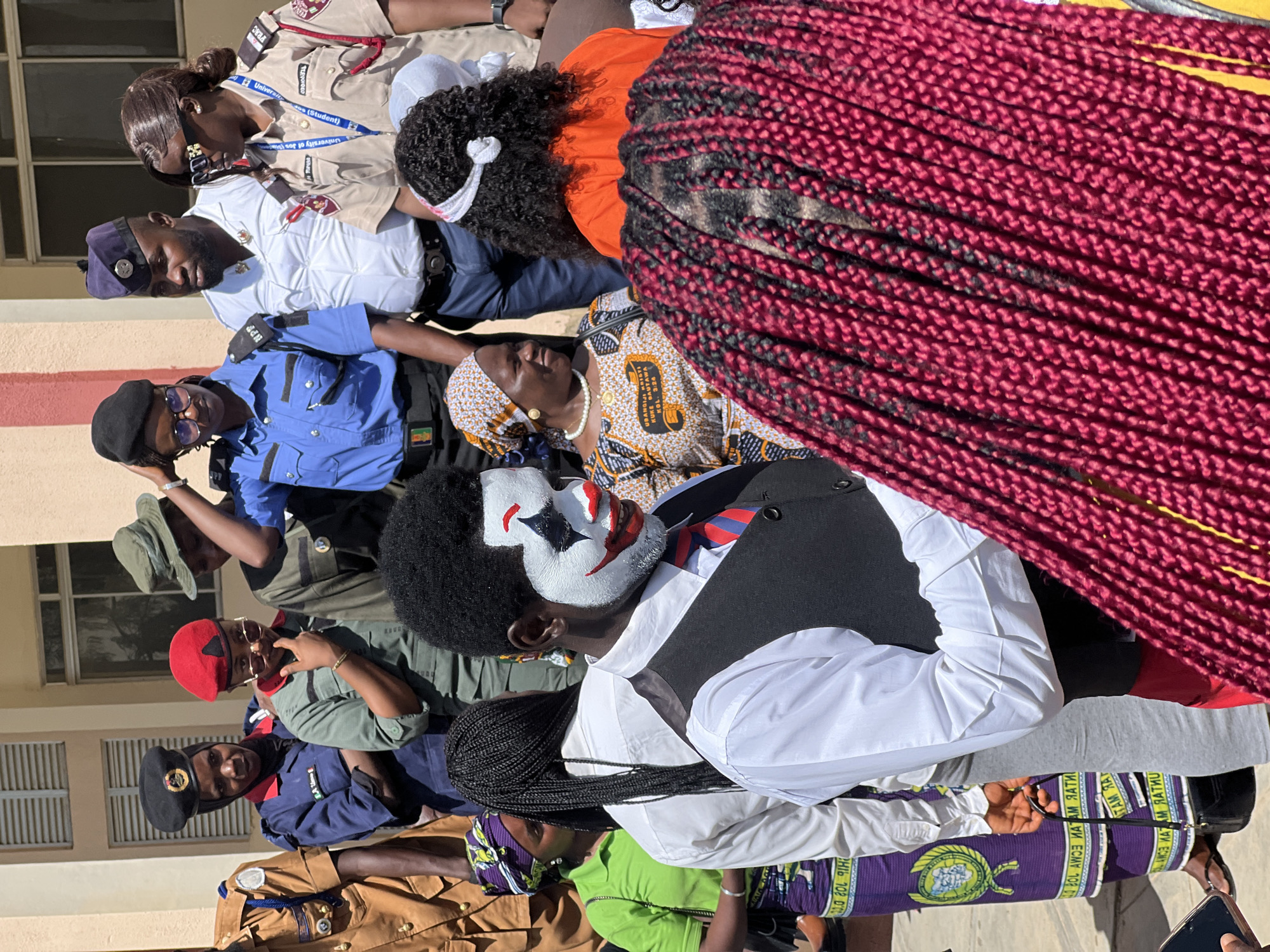 In Rafin Par, Nigeria, a man with clown makeup, characterized by white face paint, red lipstick, and black diamond-shaped eye accents, stands in the foreground. He is dressed in a black vest over a white collared shirt and a red and blue striped tie. Behind him, a group of people are gathered. To his left, a woman in a green shirt and purple and green patterned skirt is visible, her hair in long black braids. Further left, a man in a brown uniform with a patch on his chest stands. Several individuals in various uniforms, including blue, olive green, and tan, are also present, some wearing caps. One woman in a tan uniform wears a lanyard with "University of Jos" printed on it. To the right of the clown, a woman in an orange top and a white headscarf is partially visible, her curly dark hair adorned with a white band. Another woman in a colorful patterned headscarf and dress, with a pearl necklace, is looking to her left. The environment appears to be outdoors, with a building and its windows visible in the background.