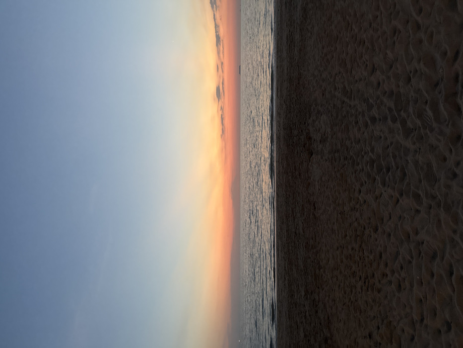 The image presents a static, panoramic view of a beach and ocean at sunset in Phra Ae, Thailand. The lower portion of the frame features dark, textured sand exhibiting numerous indentations, consistent with footprints or natural ripples. The midground is comprised of the calm, dark surface of the sea, with subtle waves visible near the immediate shoreline. On the distant left horizon, a faint light source is discernible. Towards the far right horizon, a small, dark, indistinct silhouette, consistent with a vessel or distant landmass, is present. The upper portion of the frame displays a sunset sky transitioning from a clear blue at the zenith, through a light blue and soft orange band, to a vibrant orange and reddish glow above the horizon line. Sparse, wispy clouds are present, primarily in the upper right quadrant, where a faint white point, possibly a celestial body, is also visible. No individuals, explicit actions, or direct interactions are depicted within the scene.