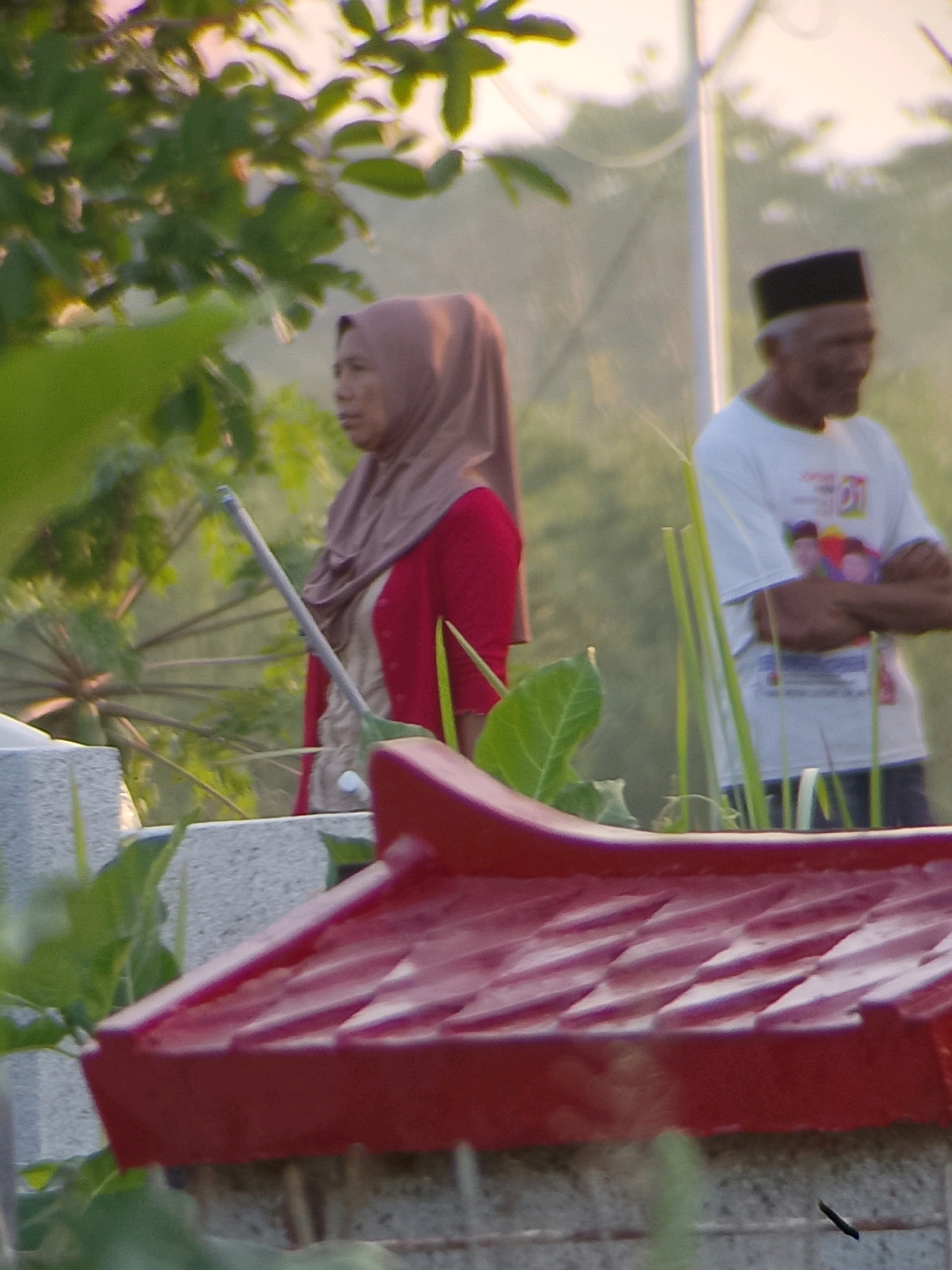 The image depicts an outdoor scene with two individuals, possibly in Jember, Indonesia. In the foreground, a red, textured object with angled surfaces, resembling a roof or decorative element, is prominently displayed. Behind this, a woman wearing a brown hijab and a red cardigan stands partially obscured by foliage. She holds a long, thin object. To the right, a man wearing a white t-shirt with a colorful graphic and a black skullcap is visible. He has his arms crossed. The background features greenery and a light-colored vertical pole. The lighting suggests it might be daytime, possibly late afternoon or early morning due to the soft light and potential golden hour tones in the upper portion of the image. The overall impression is a candid shot of people in a semi-natural or garden-like setting.