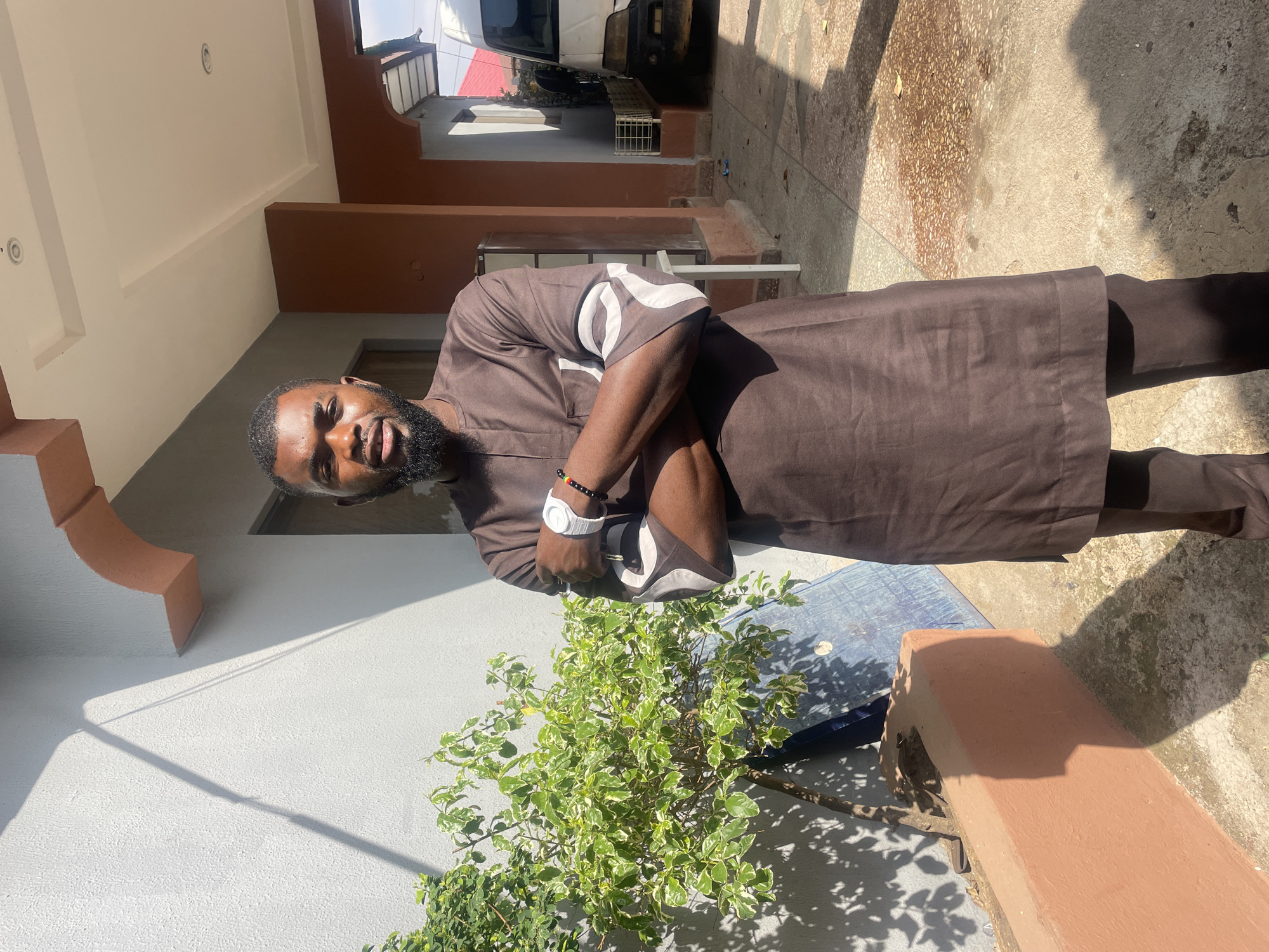 A man stands outdoors. He has a beard and wears a brown shirt and pants with white markings on the shirt's sleeves. He has a white wristwatch. He is in front of a white wall, a small green plant, and a blue object partially visible. The ground is concrete. A building and a car are visible in the background. Given the prompt, this is likely in the city of Jos, Nigeria.