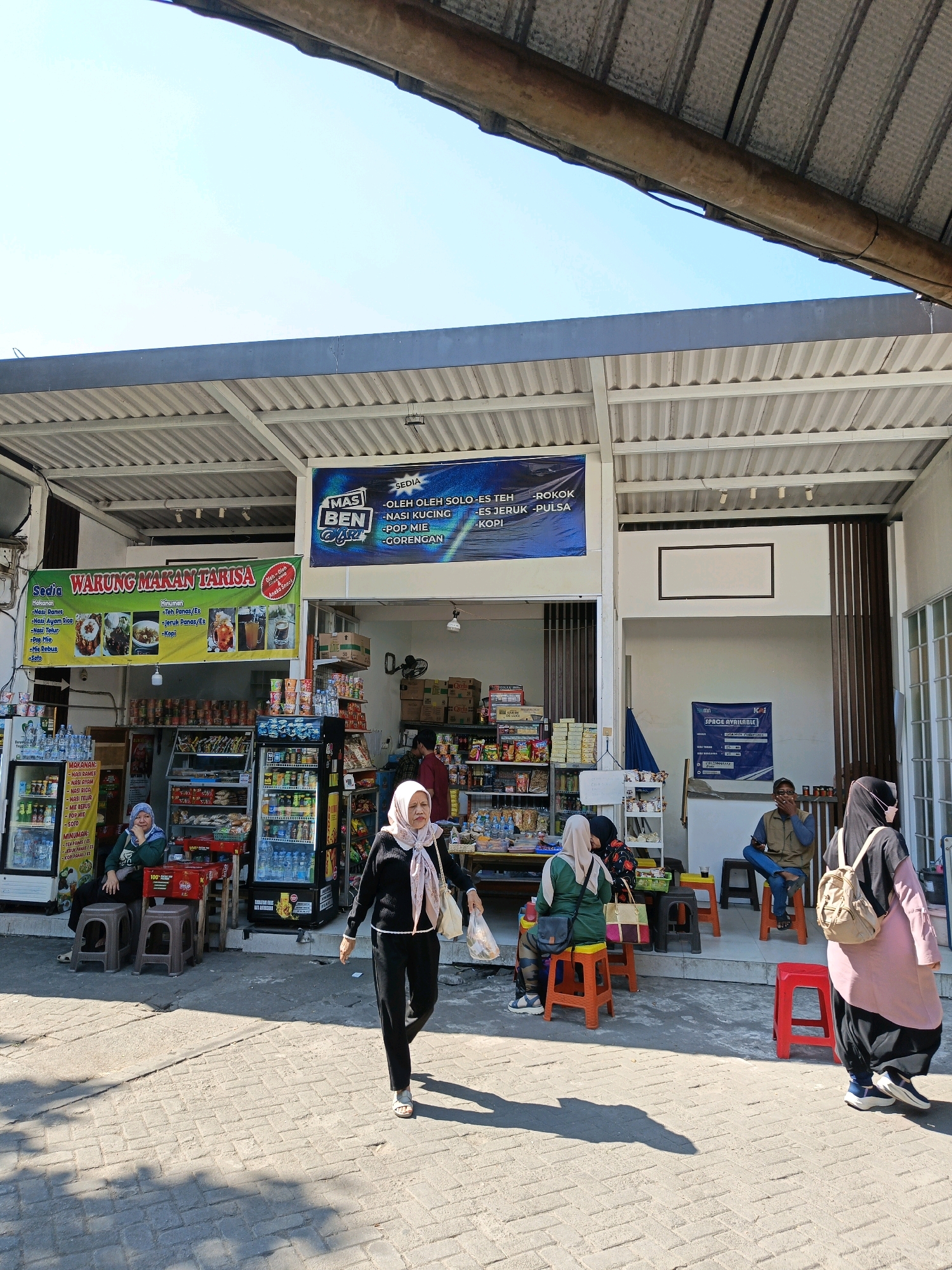 In the city of Surakarta, Indonesia, this outdoor scene captures a lively roadside stall and convenience store under a corrugated metal awning. Several individuals are present, some seated on colorful plastic stools while others are walking. A woman in a black outfit and pink hijab walks towards the viewer, carrying a plastic bag. Another woman, also in a hijab, is seated at a table. A man in a vest and jeans is seated and smoking, facing left. A third woman in a green hijab is also seated. The store itself is stocked with various food and drink items, visible through glass-fronted refrigerators and shelves. Prominent signage indicates the establishment is "WARUNG MAKAN TARISA" and also advertises "MAS BEN" with a menu of offerings like "NASI SOLO," "ES TEH," and "ES JERUK." The ground is paved with interlocking bricks, and shadows are cast across the scene by the bright sunlight.