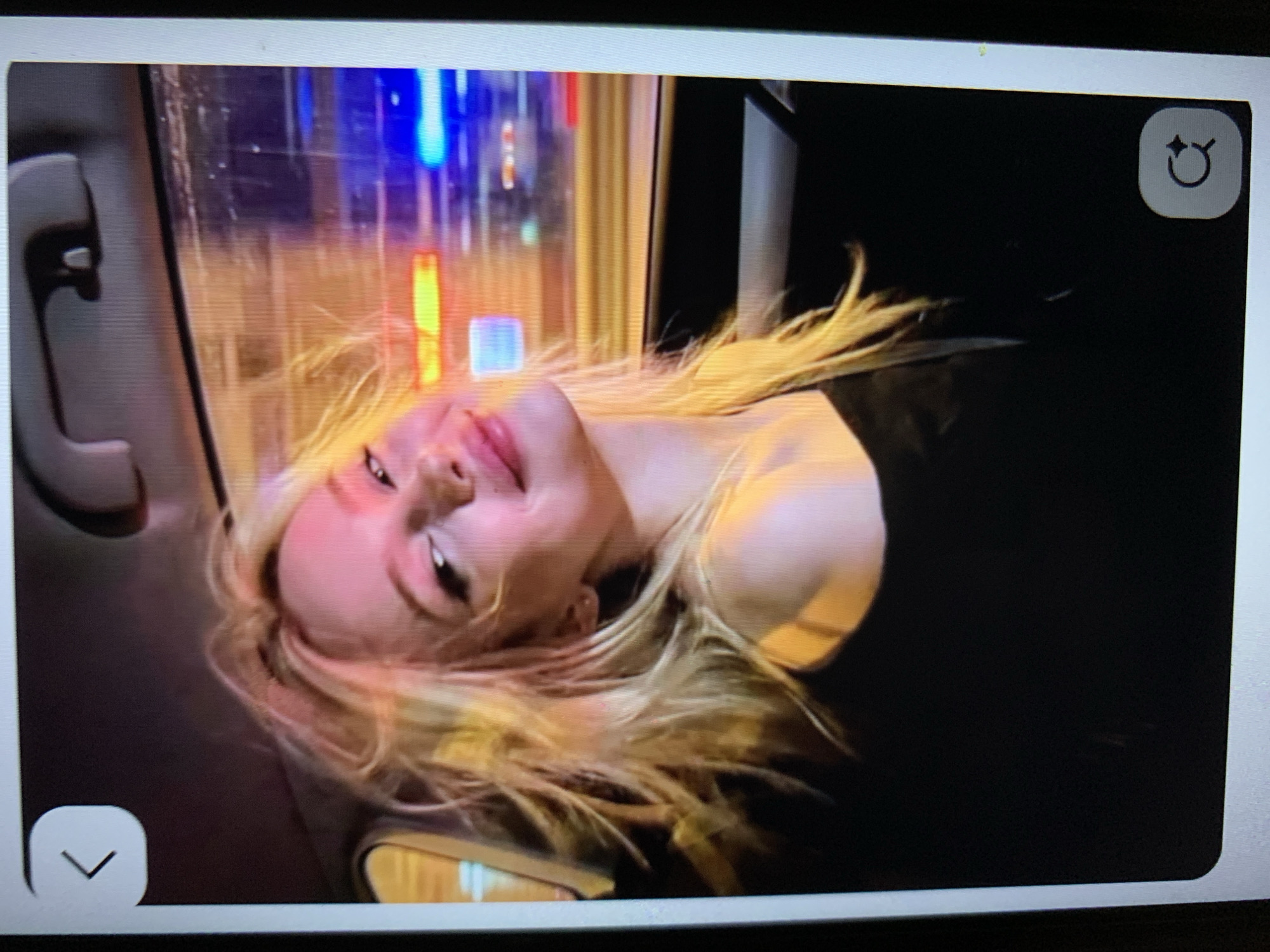 A young blonde woman with her hair styled loosely is sitting in the passenger seat of a car. The background outside the car window is blurred and illuminated by colorful lights, suggesting movement through a city at night. The woman is wearing a black off-the-shoulder top and is looking towards the camera with a slight smile. Her makeup is visible, with pink lipstick and eyeliner. The scene is captured from a slightly low angle, and the overall impression is candid and spontaneous. There is no discernible information about the location of Lymanka, Ukraine in the image.