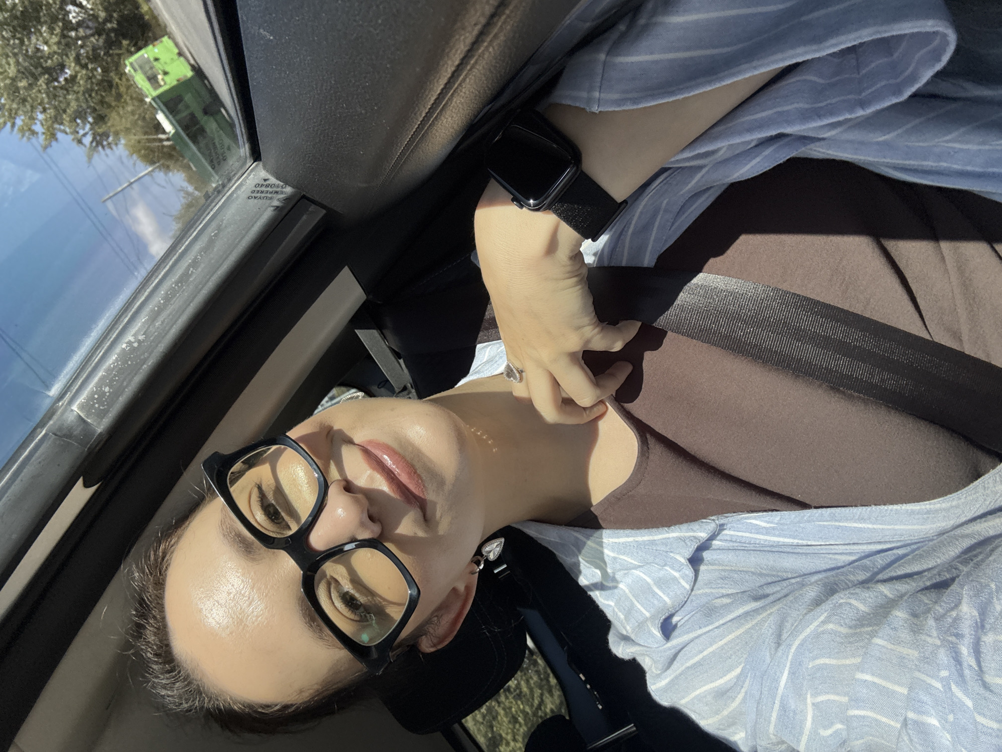 A woman is in the passenger seat of a car, wearing glasses, a blue and white striped shirt, and a brown tank top. She has a heart-shaped earring on her left ear and a ring on her right index finger. A black smartwatch is visible on her left wrist. She is wearing a seatbelt. Outside the car window, a green bus is visible on a road lined with trees and power lines. The sky is blue with some clouds. The lighting suggests it is daytime, with sunlight casting shadows. The location is not specified as Kazan, Russia.