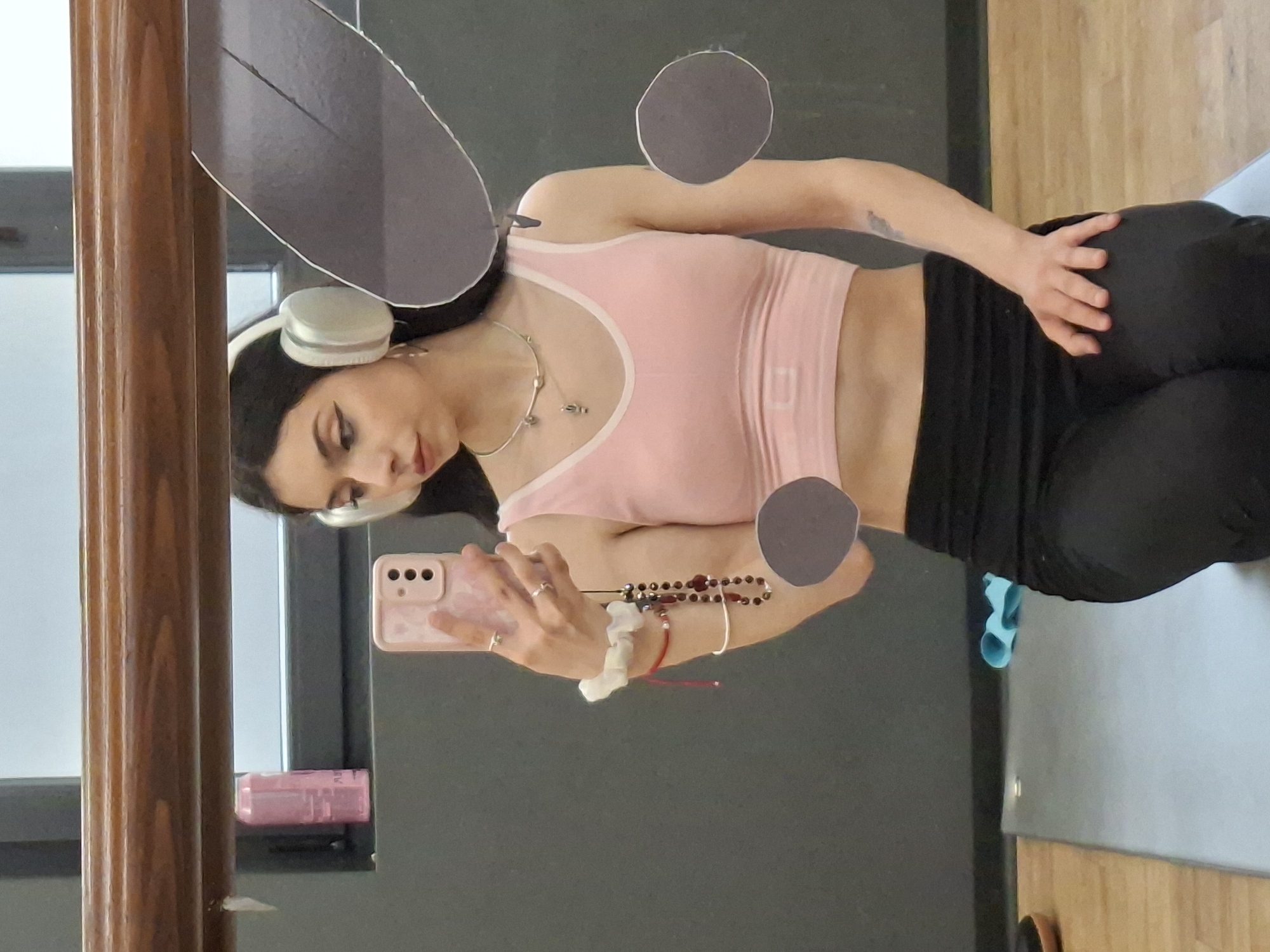 A young woman with dark hair is posing for a selfie in a mirror. She is wearing white headphones, a light pink tank top, and black leggings. She holds a pink smartphone in her hand, adorned with a white scrunchie, a red bracelet, and a beaded bracelet. Her expression is slightly angled towards the phone. The reflection in the mirror shows a glimpse of a yoga mat and wooden flooring, indicating an indoor setting. The environment outside the window suggests an urban context, possibly in Sofia, Bulgaria, indicated by the architectural style and lighting. The presence of a wooden frame and a partially visible pink bottle on a shelf add to the domestic ambiance.