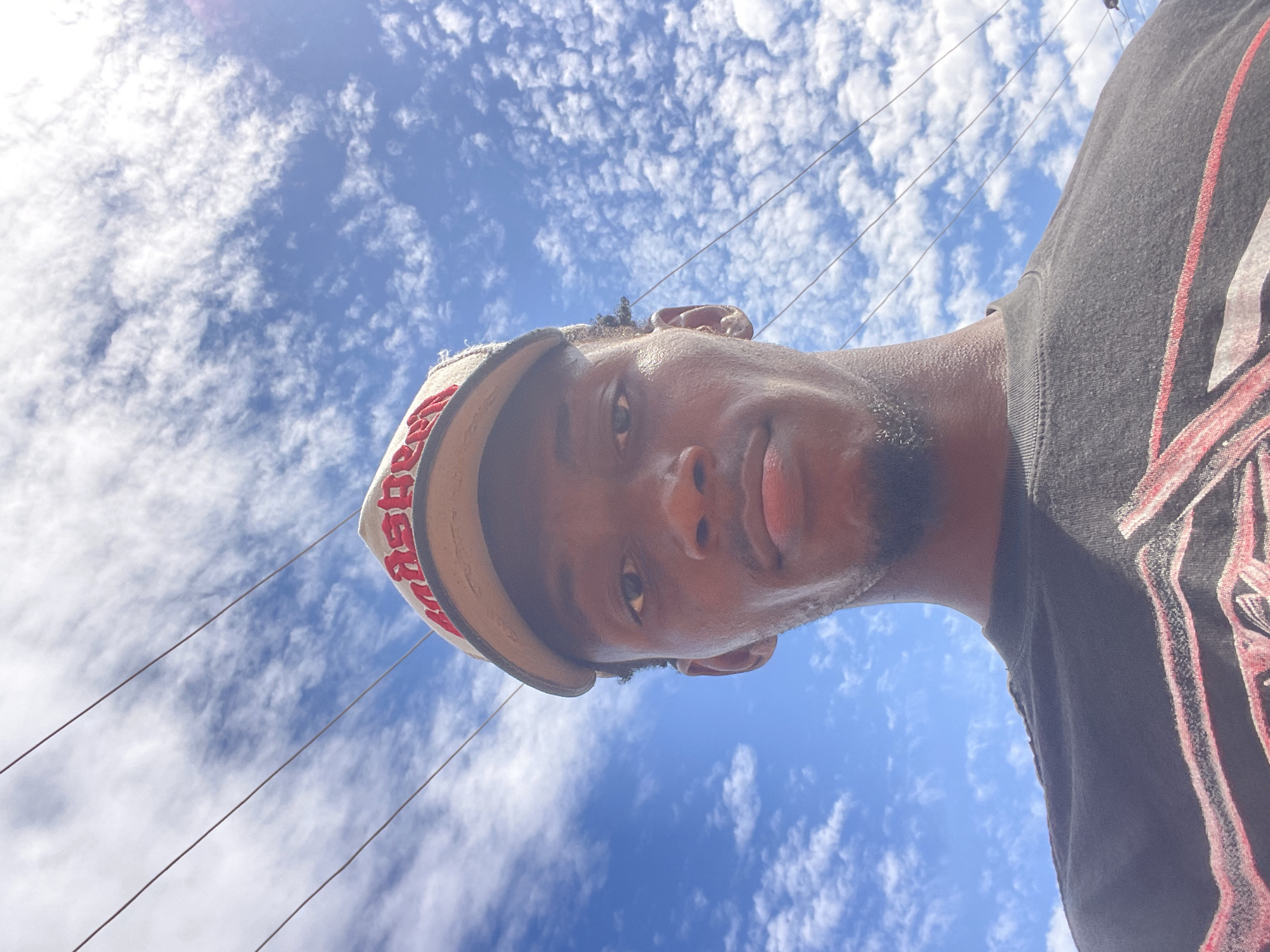 This image captures a low-angle perspective of a dark-skinned male subject, whose head and upper torso are visible on the right side of the frame. He is looking directly up towards the camera. The man wears a dark t-shirt with a red pattern or design on the chest area, and a light-colored baseball cap. The word "PREMIER" is partially visible in red, reversed lettering on the brim of his cap. His expression is neutral, and his skin appears to have a slight sheen, possibly from sweat or humidity.
The background consists of a clear, bright blue sky dotted with numerous small, fluffy white clouds, suggesting fair weather conditions. Several thin, dark power lines stretch horizontally across the upper portion of the sky, indicating an outdoor, possibly urban or semi-urban, setting. The bright illumination suggests it is daytime, likely midday. The scene takes place in Lokoja, Nigeria.