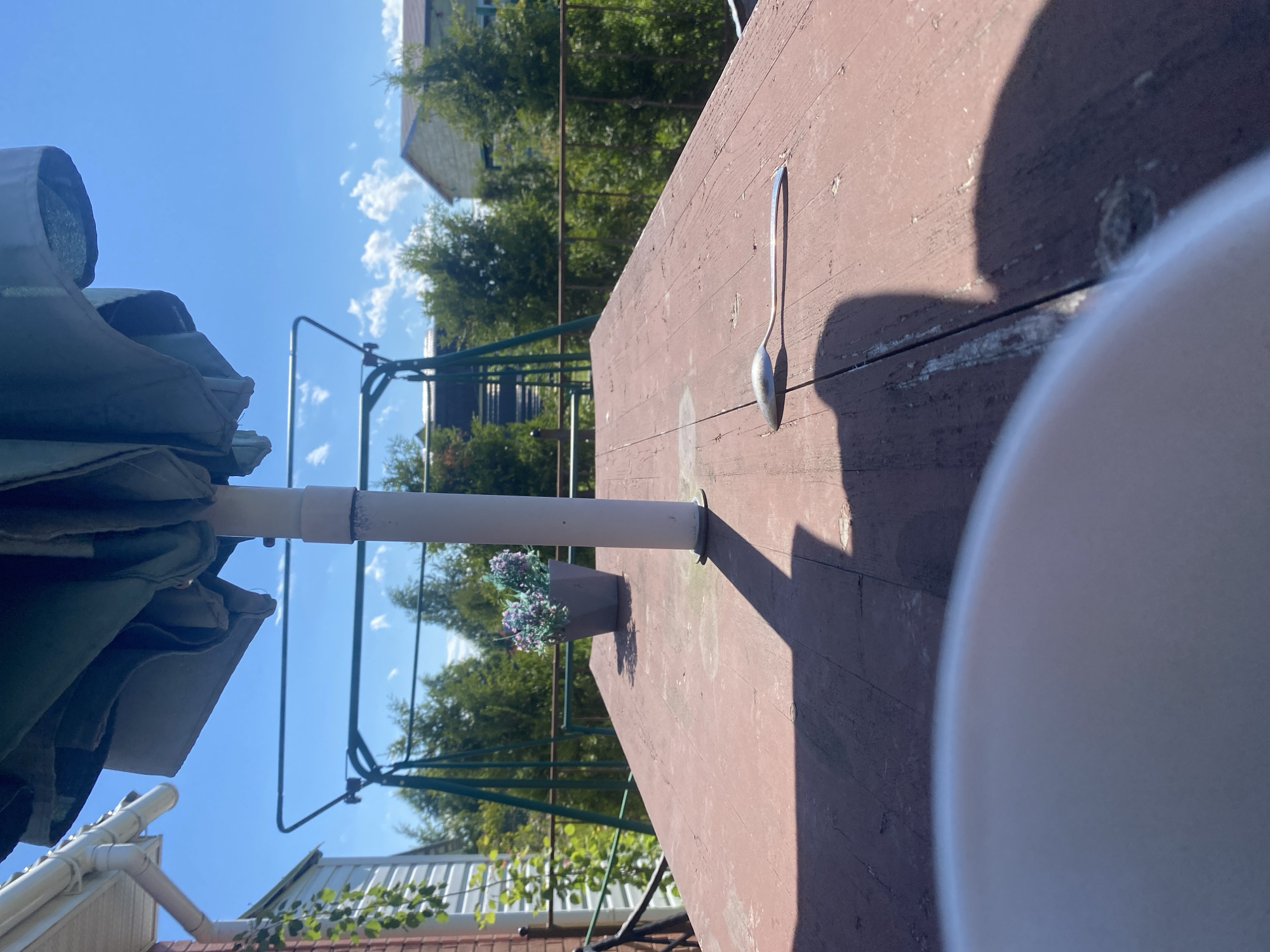 In Bol'shoe Zaborod'e, Russia, an outdoor scene unfolds under a clear blue sky with scattered white clouds. The foreground is dominated by a weathered, reddish-brown wooden table, its surface showing signs of age and use. A white umbrella pole stands vertically in the center of the table, casting a prominent shadow. To the left of the pole, a gray planter holds artificial purple and green flowers. Further to the right on the table, a single silver spoon rests, casting its own shadow. The middle ground reveals a green metal swing set. Beyond the swing set, a row of lush green trees forms a natural backdrop, with houses visible through the foliage. The scene is brightly lit by sunlight, suggesting it is daytime.