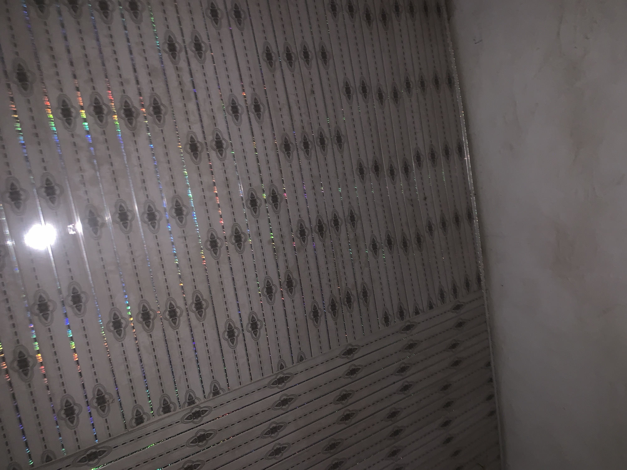 The image shows an indoor scene, likely a ceiling, with a patterned design. The ceiling is made of rectangular panels with a white base color and a repeating dark gray ornamental pattern consisting of four leaf-like shapes arranged in a diamond formation. Horizontal lines of small dashes run between the larger patterns. The ceiling panels are angled, suggesting they form a sloped or vaulted surface. Some of the panels exhibit a subtle iridescent effect, reflecting light in rainbow hues. A bright light source, possibly a bulb, is visible in the upper left quadrant of the image, causing a significant glare. The lower portion of the image shows a plain, off-white or light gray wall that meets the ceiling at an angle. There are no people or identifiable objects present, and no context is provided to determine the specific location within Jalingo, Nigeria, other than it is an interior space.