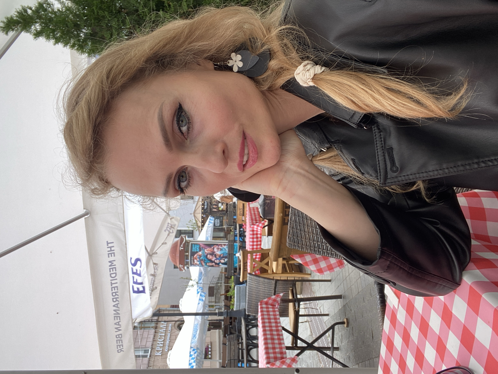 This is a selfie taken outdoors at a cafe in Minsk, Belarus. The woman in the foreground is light-skinned with blonde hair styled in two pigtails and is wearing a black leather jacket and large floral earrings. Her left hand is resting on her cheek, and she is looking at the camera. Behind her, tables with red and white checkered tablecloths are set up. Umbrellas, some with the Efes logo, provide shade. In the background, there are buildings, trees, and an advertisement poster. The overall scene suggests a relaxed outdoor dining environment.