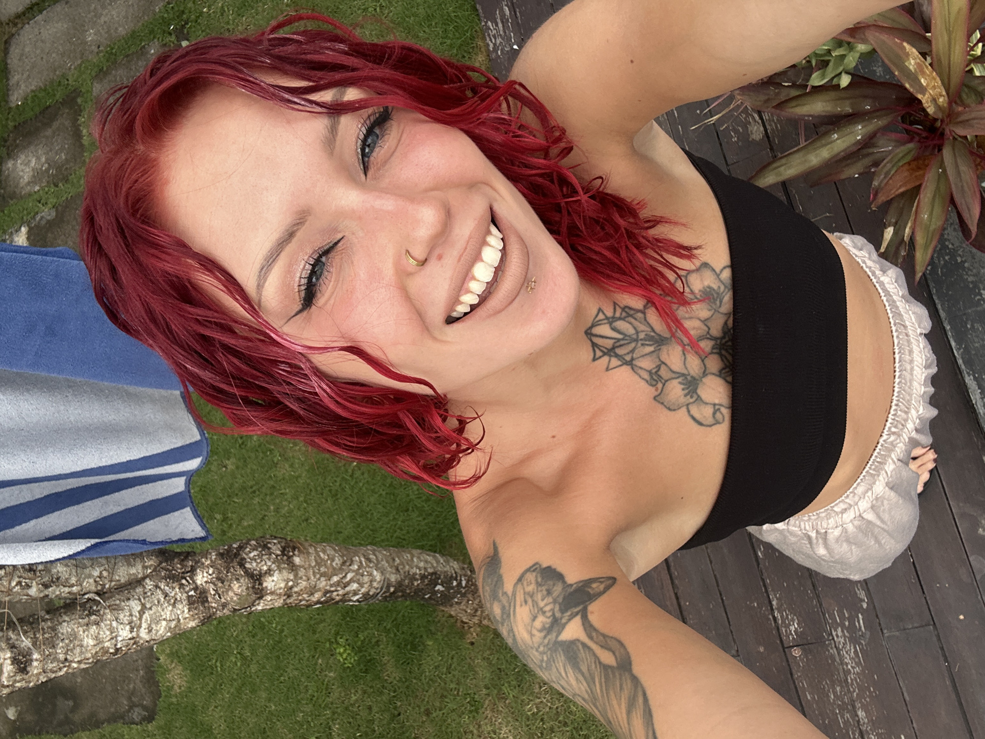 A vibrant, high-angle selfie captures a young woman with bright red, wet hair, light eyes, and a wide, beaming smile. Her eyes are adorned with dark eyeliner and mascara. She has a gold-colored hoop piercing on her nose and a small gold stud on her philtrum. Visible tattoos include a large floral design on her upper chest and shoulder, and a stylized animal (possibly a bird or dog) on her right upper arm.
She is dressed in a black crop top or sports bra and light-colored, possibly white or light grey, shorts with a ruffled elastic waistband. Her wet hair suggests she may have recently been swimming or showering.
The setting is an outdoor area, likely a patio or backyard in North Kuta, Indonesia, evident from the tropical flora and natural elements. She is positioned above a weathered wooden deck. To her left, a blue and white striped towel is draped over a tree trunk, with lush green grass visible below. A potted plant with reddish-green leaves is in the background to her right. The scene is illuminated by natural daylight, conveying a cheerful and relaxed atmosphere. There is no visible text in the image.