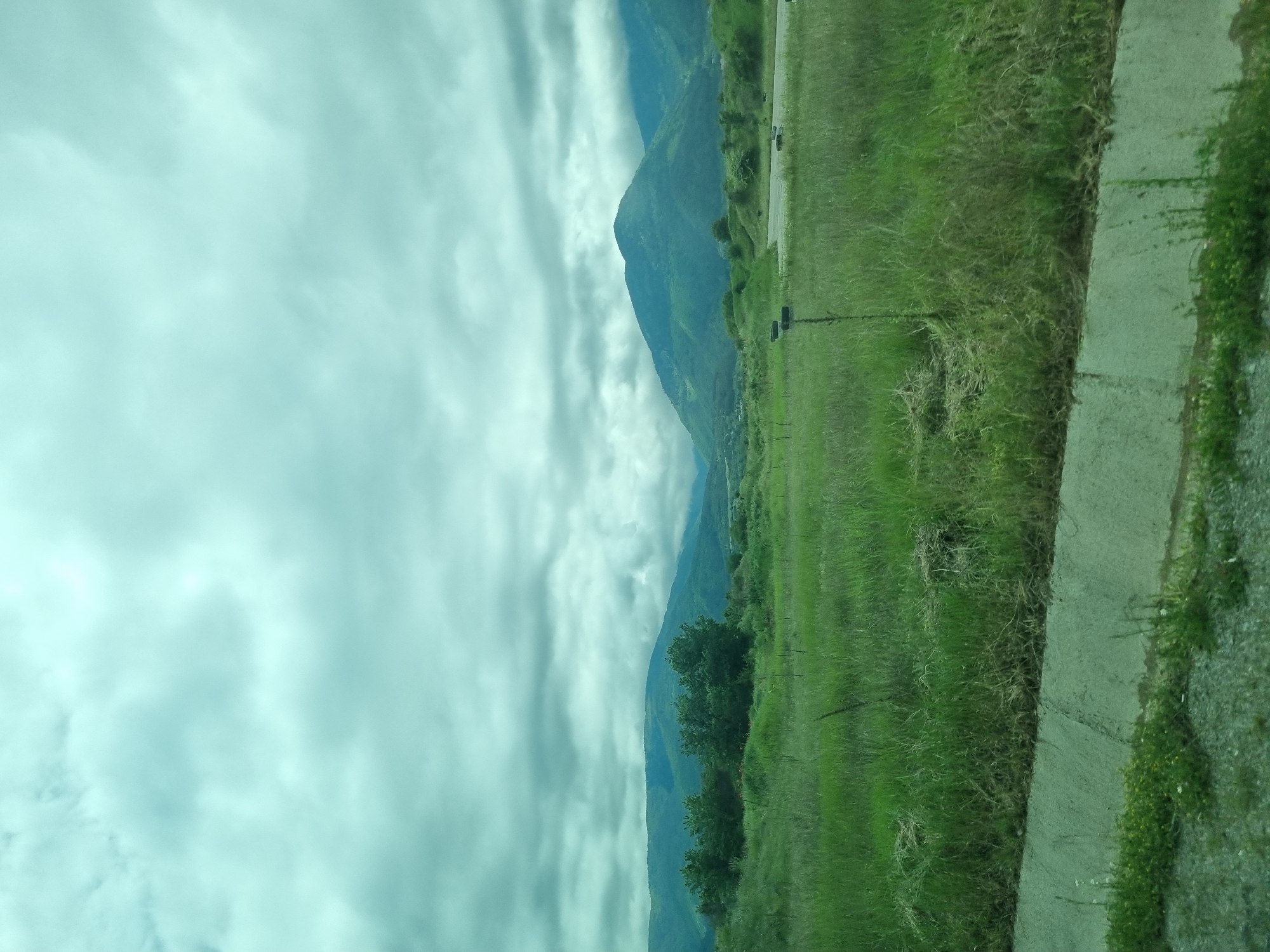 The image depicts a landscape scene, likely taken from a moving vehicle. On the right, a strip of green grassy land with a concrete sidewalk. Adjacent to this is a vast expanse of green field. In the distance, there are mountains partially obscured by low-hanging, overcast clouds that dominate the sky. The photograph is taken in Yahinovo, Bulgaria.