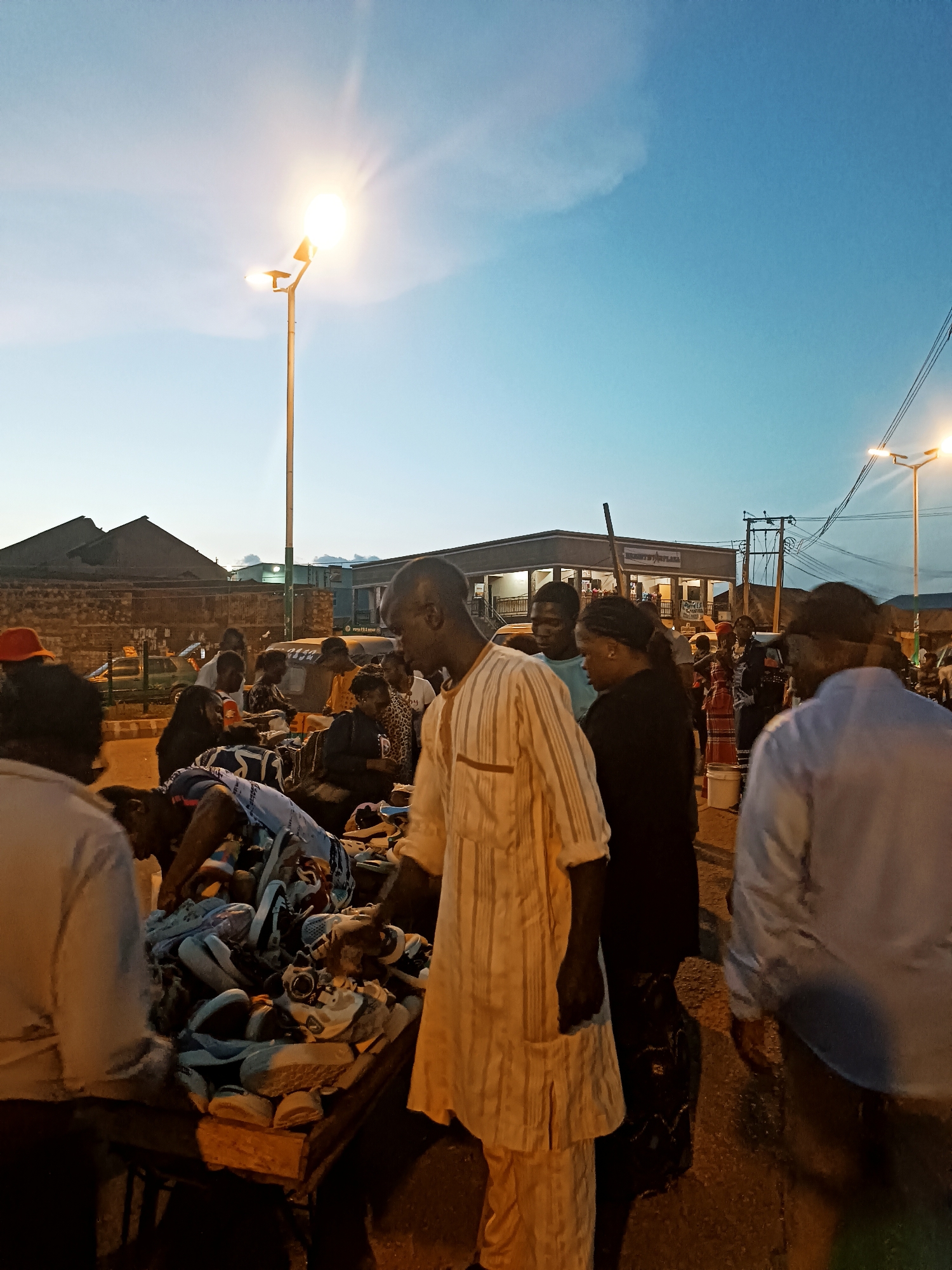 This image captures an outdoor market scene at dusk or early night in Unknown, Unknown. The sky transitions from a light blue at the horizon to a deeper blue overhead, with two prominent streetlights illuminating the area. The leftmost streetlight, mounted on a tall pole, emits a powerful glare with distinct light rays, indicating its brightness.
Approximately 15 to 20 individuals, primarily adults, are present in the frame. In the center-left foreground, a man in a light-colored, vertically striped traditional tunic and trousers stands, looking down at a large, disorganized pile of assorted footwear, including sneakers and sandals, displayed on a wooden cart or table. Adjacent to him, a woman in a dark top also faces the merchandise. In the far right foreground, a man in a light blue long-sleeved shirt is partially visible, walking away from the viewer. Other individuals are dispersed in the mid-ground, some gathered around additional market stalls, appearing to browse or interact.
In the background, a dark-roofed building is visible on the left. Toward the center-right, a larger, multi-story commercial building features several windows and some indistinct signage, with partial text "MONASTO MAYFLA..." and "FURNI..." discernable on separate signs. Overhead, a dense network of black electrical wires extends across the upper right quadrant, connected to utility poles. The ground appears to be an unpaved surface or dark asphalt. The overall environment suggests a bustling commercial activity during the twilight hours.