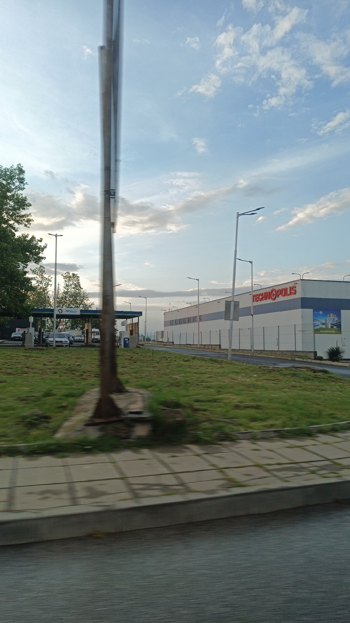 The image shows a view from a moving vehicle, with a blurred metal structure obstructing part of the view. On the right is a "TECHNOPOLIS" store, a large building with white and blue sections and advertising on its facade. A fence runs along the perimeter. Streetlights line the road.
To the left of the store is a gas station identified as "TOP Wash" with several cars parked under the canopy. The foreground consists of a strip of grassy area and a concrete sidewalk, followed by what appears to be a road or a surface reflecting light, likely the vehicle's window or dashboard. The sky is partly cloudy, suggesting daylight.
The location is likely in Burgas, Bulgaria, given the presence of the "TECHNOPOLIS" store, a known retail chain in Bulgaria, and the possible presence of a TOP Wash.