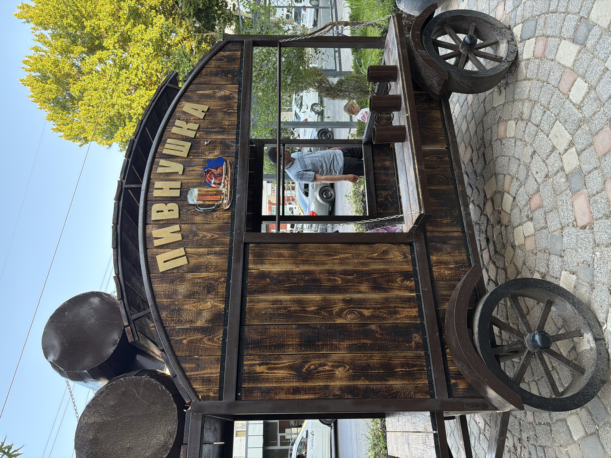 In Chirchik, Uzbekistan, a wooden cart with a rustic design resembling barrels is parked on a cobblestone pavement. The cart is adorned with the Cyrillic inscription "ПИВНУШКА" in gold lettering, accompanied by a graphic of a beer mug and food. Inside the cart, a man in a striped shirt and dark pants is visible, as well as a young child. The cart has large wooden wheels and is situated outdoors, with a street and buildings in the background. The scene is illuminated by natural daylight, suggesting it is daytime.