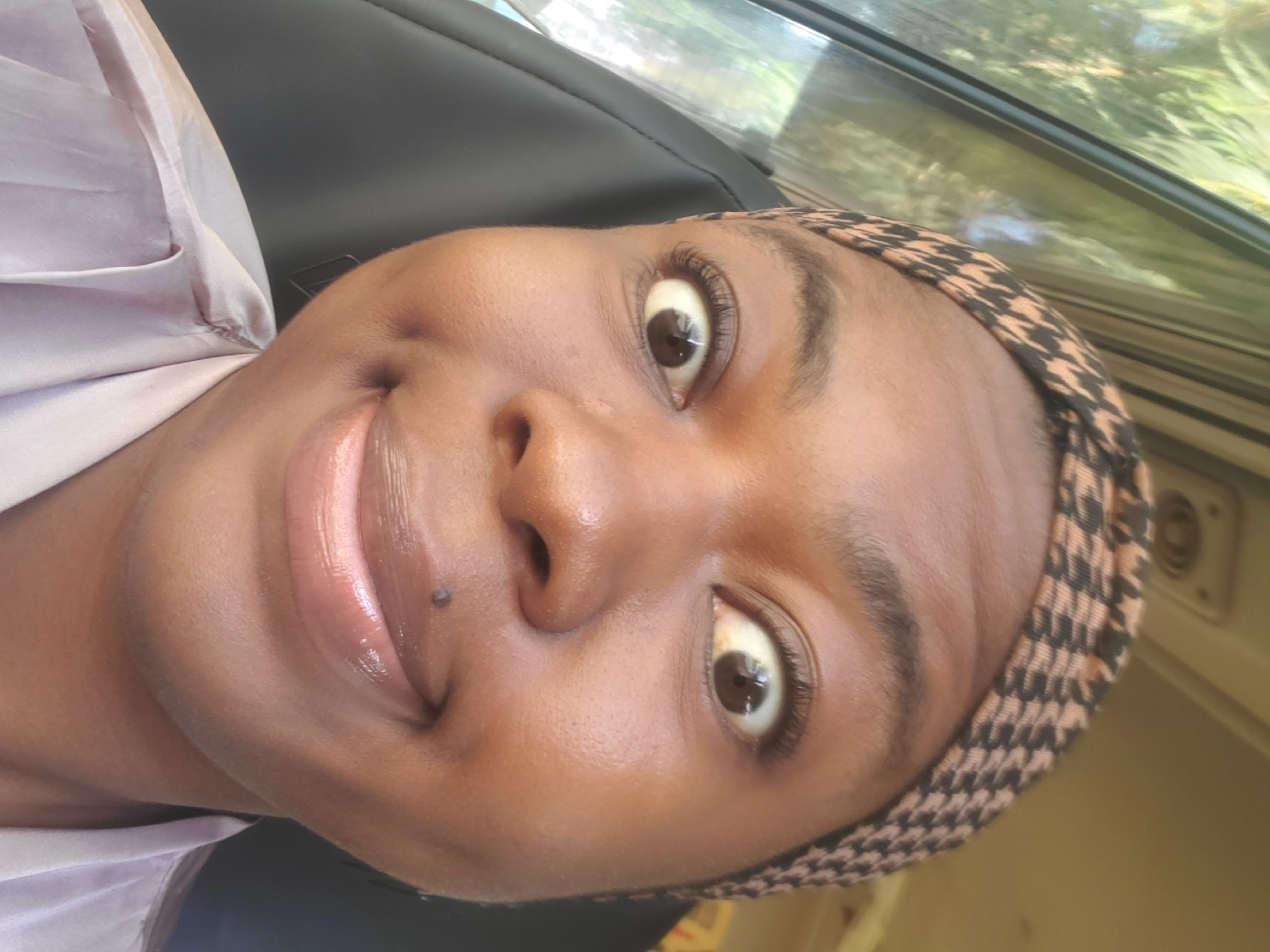 A close-up, selfie-style photograph shows a Black woman with a wide, surprised expression, her eyes looking directly at the camera. She is wearing a headscarf with a black and tan houndstooth pattern and a light-colored, silky top. A small mole is visible on her upper lip, just above the center. The background shows a car interior, with a dark seat on the right and a window with greenery on the left, suggesting the photo was taken while traveling. The lighting appears to be natural daylight. The image is consistent with a personal photograph taken in Gwagwada, Nigeria, a location known for its residential and semi-urban characteristics.