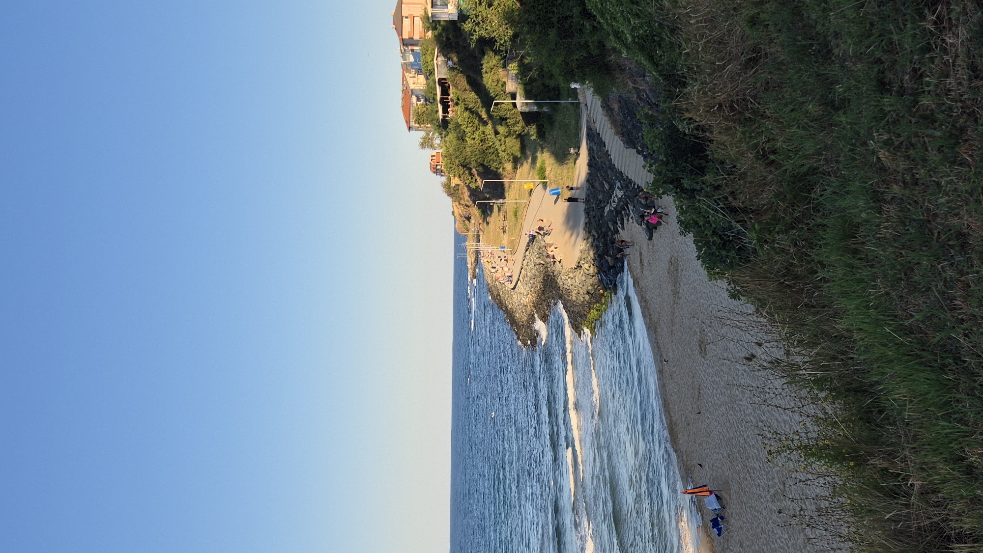 The image depicts a sunny day at a coastal area, likely the beach in Primorsko, Bulgaria. The scene showcases a tranquil seascape with gentle waves rolling onto a sandy beach. A paved walkway runs along the rocky coastline, where several people are seen strolling and enjoying the view. Some individuals are also visible on the beach, lounging on chairs or walking. The background features lush green vegetation and residential buildings situated on a hill overlooking the sea. The clear blue sky suggests pleasant weather conditions, typical of a summer day.