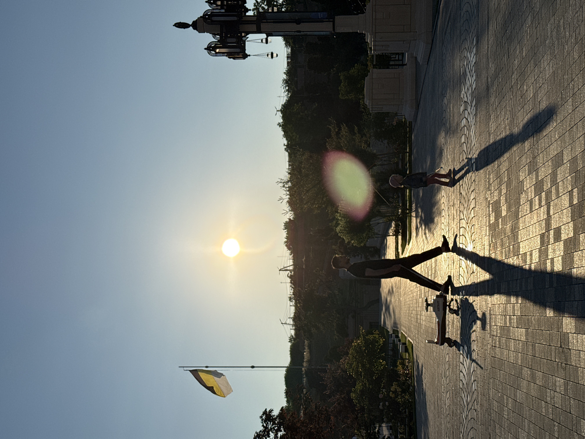 In an outdoor plaza paved with interlocking stones, a man walks from left to right across the frame, silhouetted against a bright sun near the horizon. He is pulling a child's ride-on toy, which casts a long shadow. To the right of the man, a small child stands facing away from the viewer, also silhouetted, holding a stick. In the background, a flagpole with a yellow and black flag is visible on the left, and a decorative lamppost stands on the right. The setting sun casts a warm glow on the scene, creating strong shadows and a hazy atmosphere. Trees and a gentle slope are visible behind the plaza, with a clear sky above.