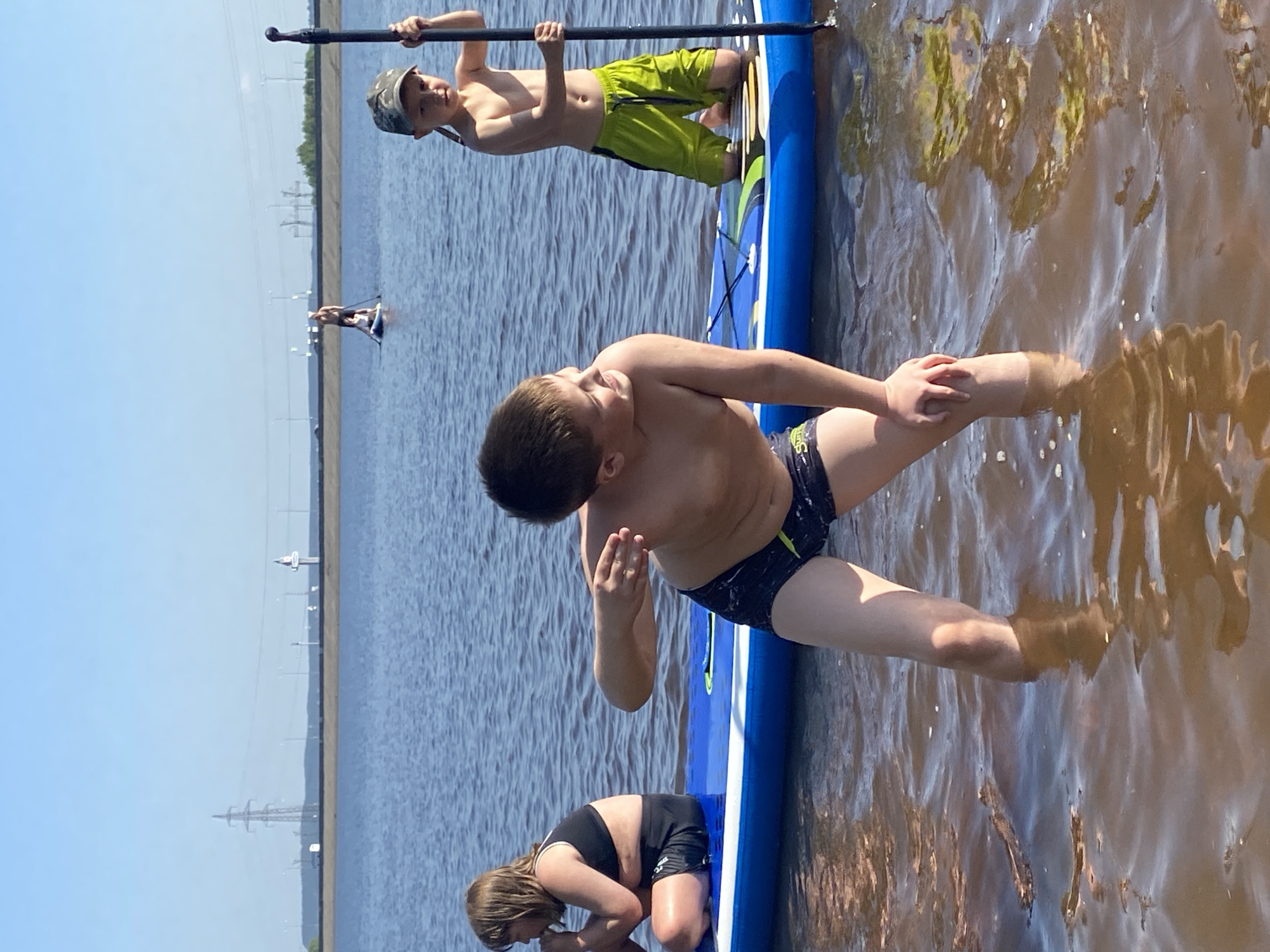 Three children are on a paddleboard in the water. One child is standing on the paddleboard at the top of the image, holding a paddle overhead. Another child is kneeling on the paddleboard at the bottom of the image. The third child is in the center of the image, partially in the water and on the paddleboard. The water is brown. There are power lines on the opposite shore. The location is Chaikovsky, Russia.