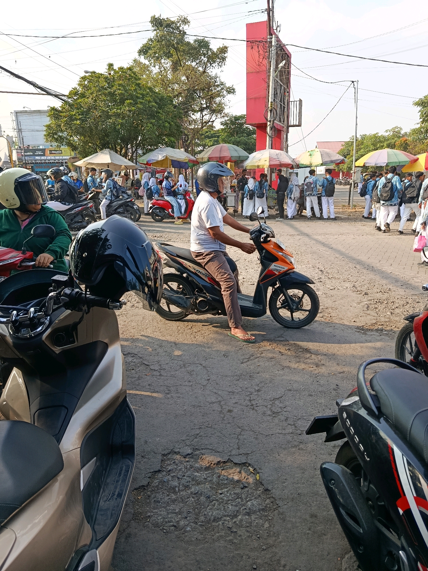 The image captures a street scene in Bululawang, Indonesia, under a bright, clear sky. In the foreground, a tan scooter is parked, with a black helmet resting on its handlebars. To the right, another scooter, primarily black with red and white accents, is also visible.
The midground is populated by several individuals and a motorcycle. A man wearing a white t-shirt, brown pants, and flip-flops is seated on an orange and black motorcycle. He is looking towards the left. Behind him, a group of students, many wearing blue uniforms and carrying backpacks, are gathered around food stalls shaded by colorful umbrellas. Some students are standing, while others are walking.
In the background, a large red structure, possibly an advertisement billboard, is prominent. Trees with lush green foliage line the street. Power lines are strung across the sky, indicating an urban or semi-urban environment. The ground is a mixture of asphalt and dirt, with some areas appearing worn and cracked. The overall atmosphere suggests a busy marketplace or school vicinity, likely during the day given the lighting conditions.