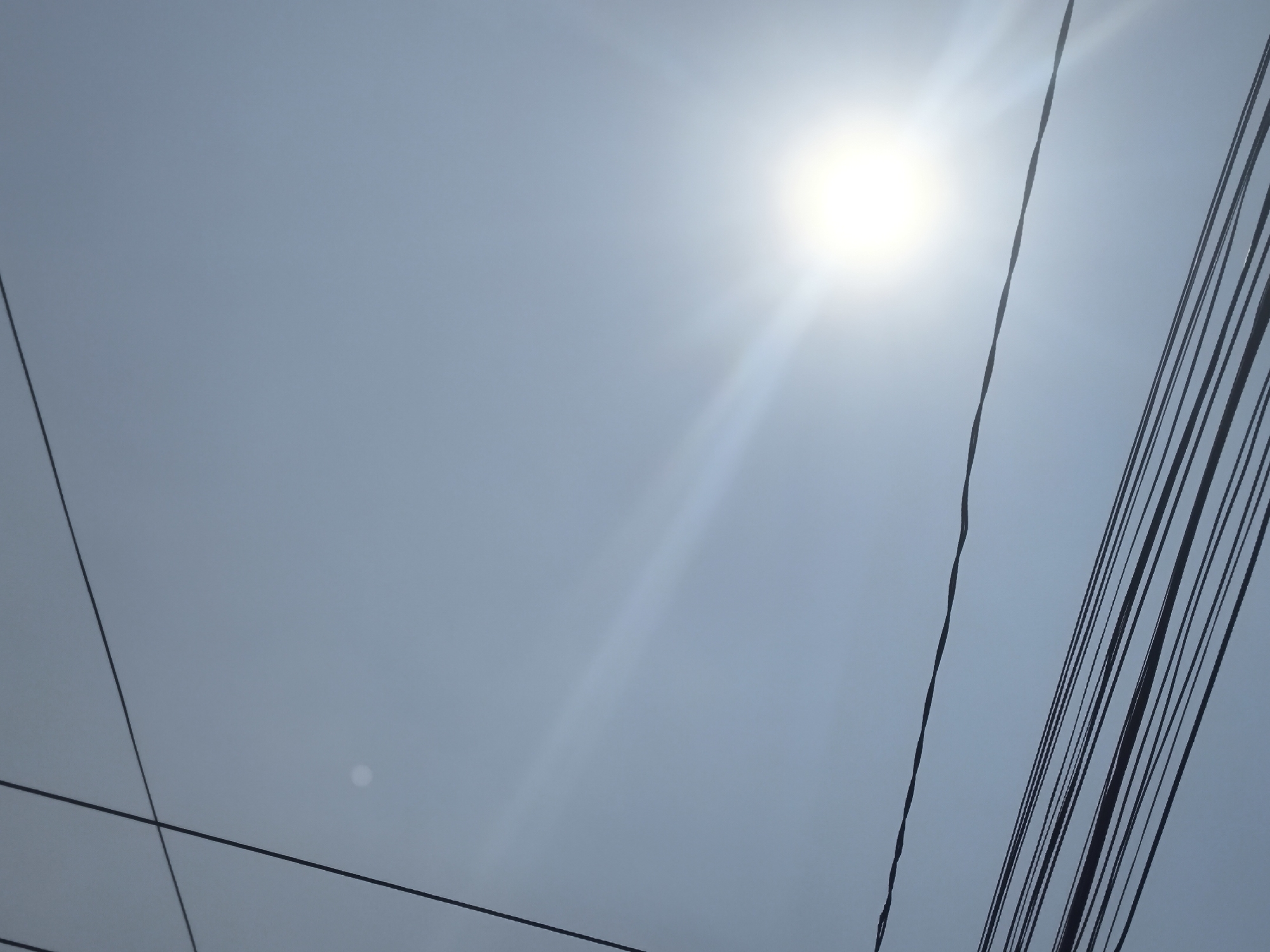 The image shows a bright sun with visible rays, casting a glare in a hazy, light blue sky. Several black electrical wires crisscross the frame at different angles, some appearing taut and others slightly slack. One cluster of wires runs diagonally from the bottom right corner towards the upper left. Another single wire runs diagonally from the top left towards the bottom right. In the background, a faint circular light or reflection is visible in the sky. The scene is likely an outdoor shot, possibly taken from a street level or elevated perspective, capturing the urban infrastructure against the sky. The overall impression is one of a typical day in an urban environment, characterized by overhead wiring and bright sunlight.