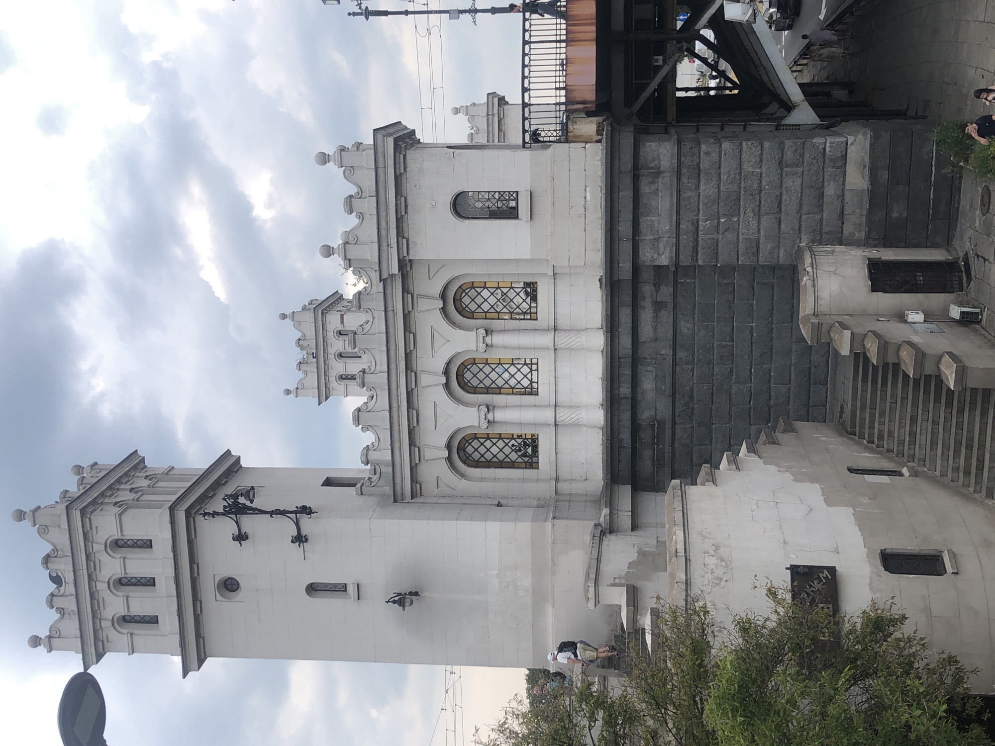 The image shows the Citadel in Warsaw, Poland, a historic fortress complex. The perspective is from an elevated position looking down and across the structure. In the foreground, a white tower with crenellations and arched windows is prominent, with a man walking up stairs to its left. Adjacent to the tower is a building with similar architectural features and more arched windows with decorative grilles. A metal bridge with railings extends to the right of this building, and a person is walking across it. The buildings are constructed of white stone and sit on a dark, rough-hewn stone foundation. Below the foundation, visible through the structure of a bridge, is a street with cars and pedestrians. The sky is overcast with gray clouds.