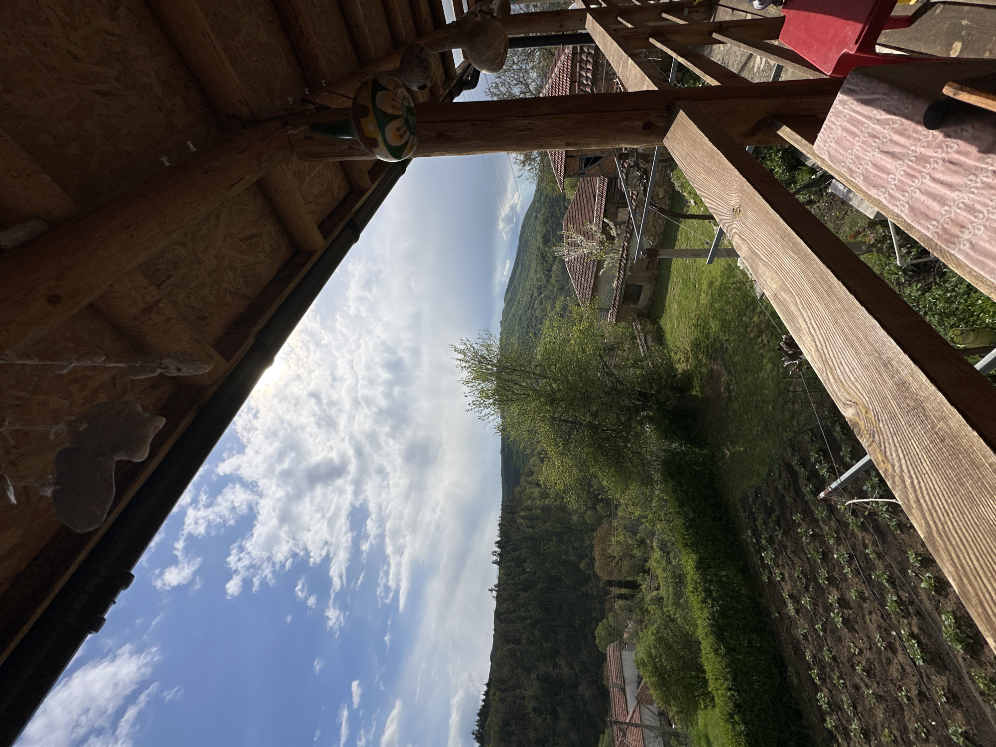 The image, taken in what appears to be Mrachenik, Bulgaria, shows a view from under a wooden structure, possibly a porch or shed. The sky is partly cloudy with blue patches. In the distance, there's a green, wooded hillside. Below the hillside, there are buildings with reddish-brown tile roofs, typical of the region. A cultivated garden is visible in the foreground. The picture is taken at an angle, making the scene appear slightly rotated.